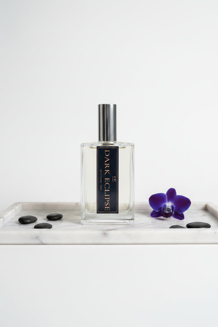"Dark Eclipse Eau de Parfum glass bottle with silver cap and black label, styled on a marble tray with black stones and a purple orchid on a white background — luxury perfume by SunShine Scents Candle Co."