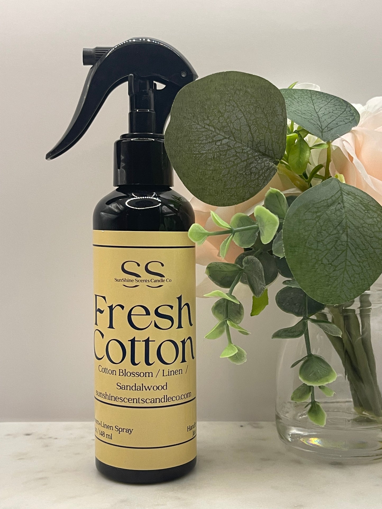 A bottle of Fresh Cotton scent room spray with a black trigger sprayer and a beige label, shown next to green eucalyptus leaves and a glass vase on a light surface.