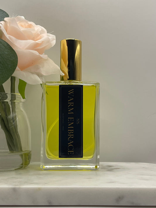 A rectangular glass perfume bottle with a gold cap and a black label reading 'WARM EMBRACE' in gold lettering, filled with yellow-green liquid. The bottle is styled with a pale peach rose and green leaves against a light gray background.
