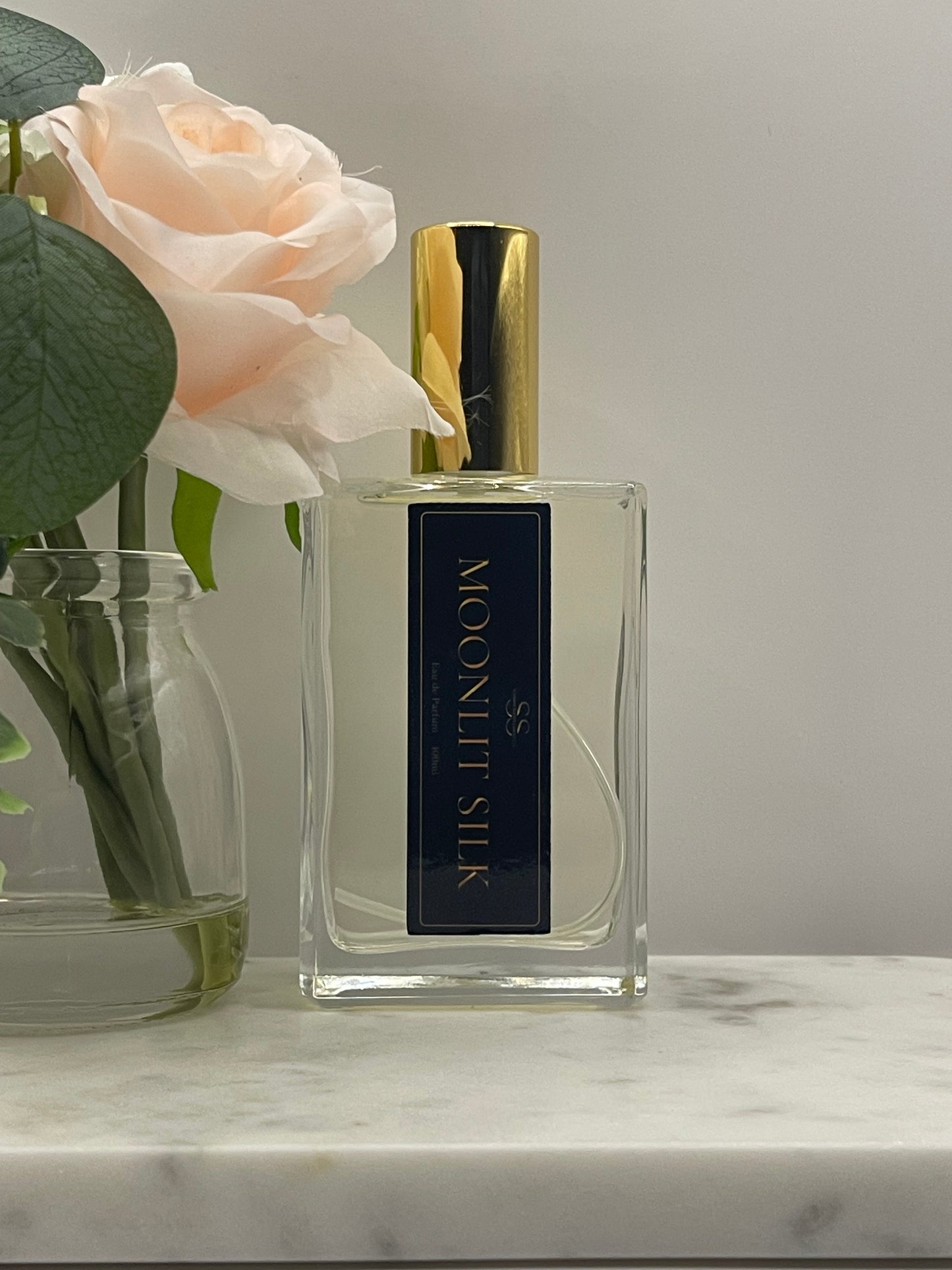 A rectangular glass perfume bottle labeled 'MOONLIT SILK' with a gold cap, shown beside a pale blush rose and green leaves against a light gray background.