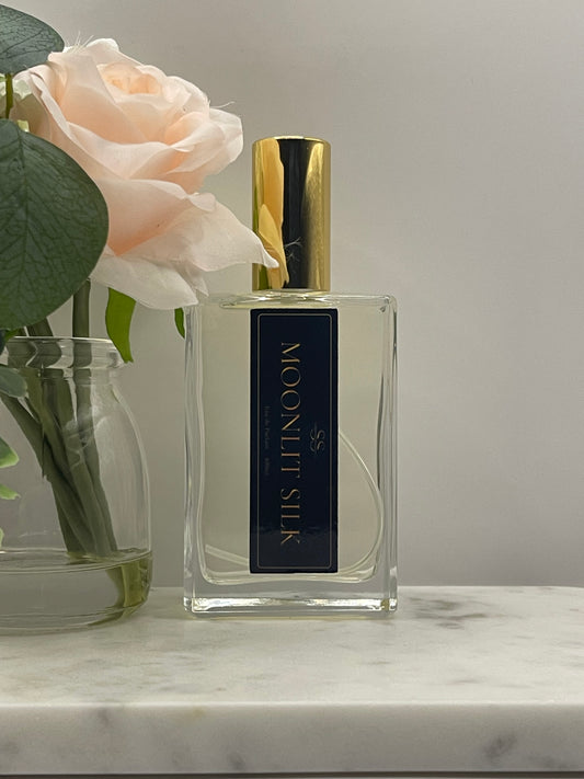 A rectangular glass perfume bottle labeled 'MOONLIT SILK' with a gold cap, shown beside a pale blush rose and green leaves against a light gray background.