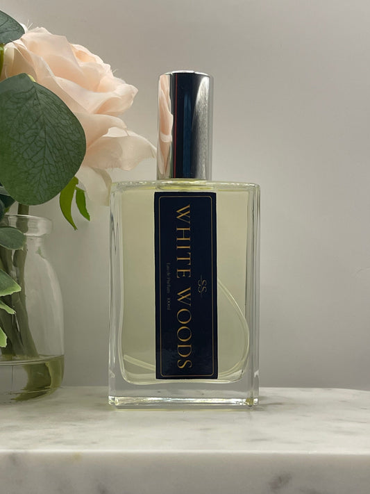 A rectangular glass perfume bottle labeled 'WHITE WOODS' in gold lettering on a dark navy label, with a reflective silver cap. The bottle is styled with pale pink roses and green leaves against a light gray background and a white marble surface.