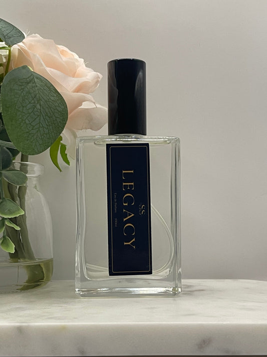 A rectangular clear glass perfume bottle with a black cap and a black label featuring gold lettering that reads 'SS LEGACY'. The bottle is styled with a pale peach rose and green leaves against a light gray background.