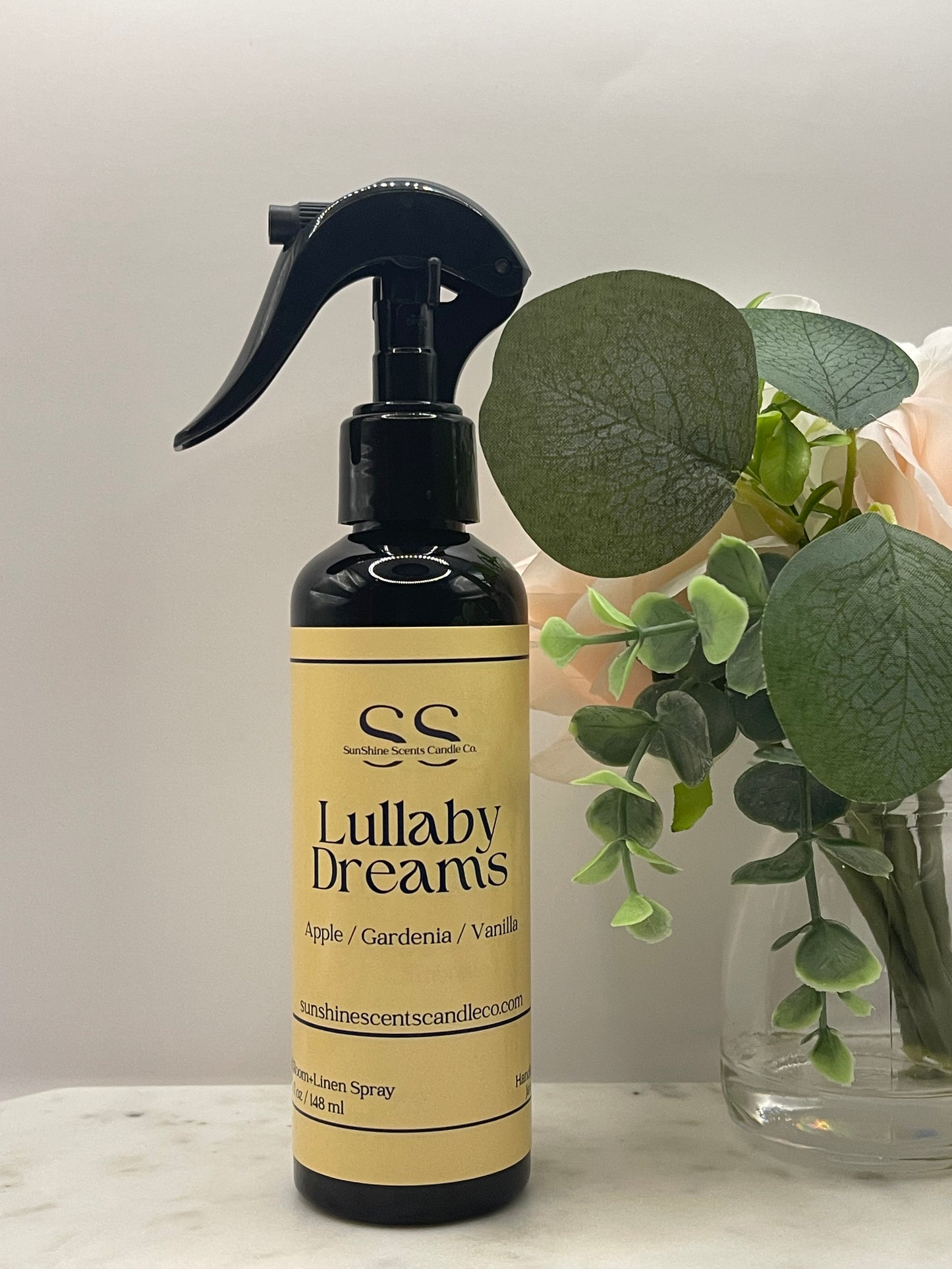 A bottle of 'Lullaby Dreams' hair styling spray by 'Gentle Secrets' with a black trigger sprayer and a beige label, styled on a white marble surface next to a vase of green leaves and pale pink roses.