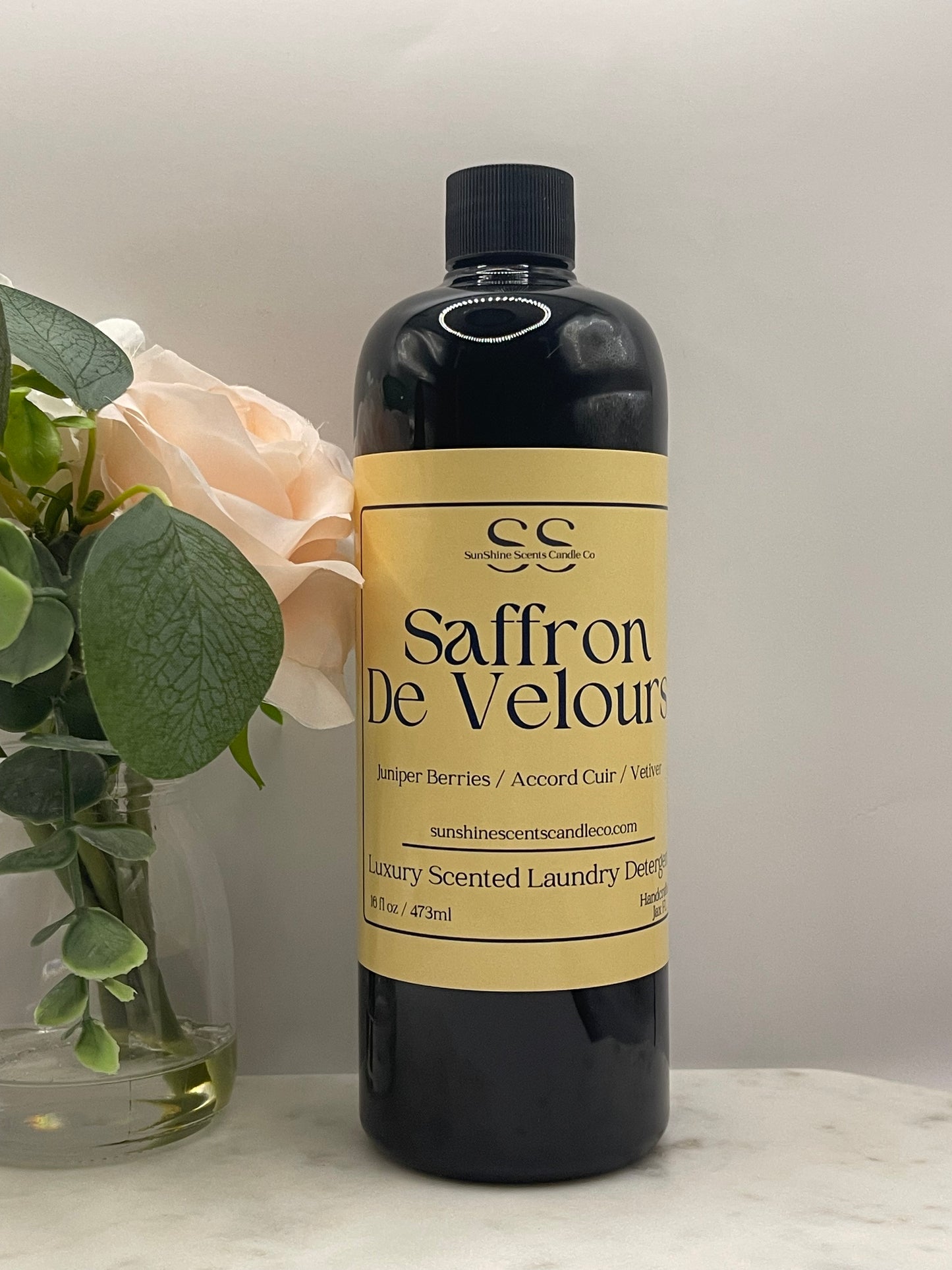 A bottle of Saffron De Veloutier luxury scented laundry detergent resting on a white marble surface next to a pale peach rose and green leaves.