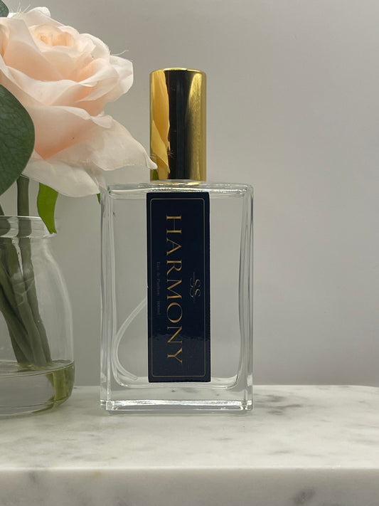 A rectangular clear glass perfume bottle with a shiny gold cap and a dark navy label reading 'HARMONY' in gold lettering. The bottle is styled with a pale pink rose and green leaves against a light gray background with a marble surface visible on the right.