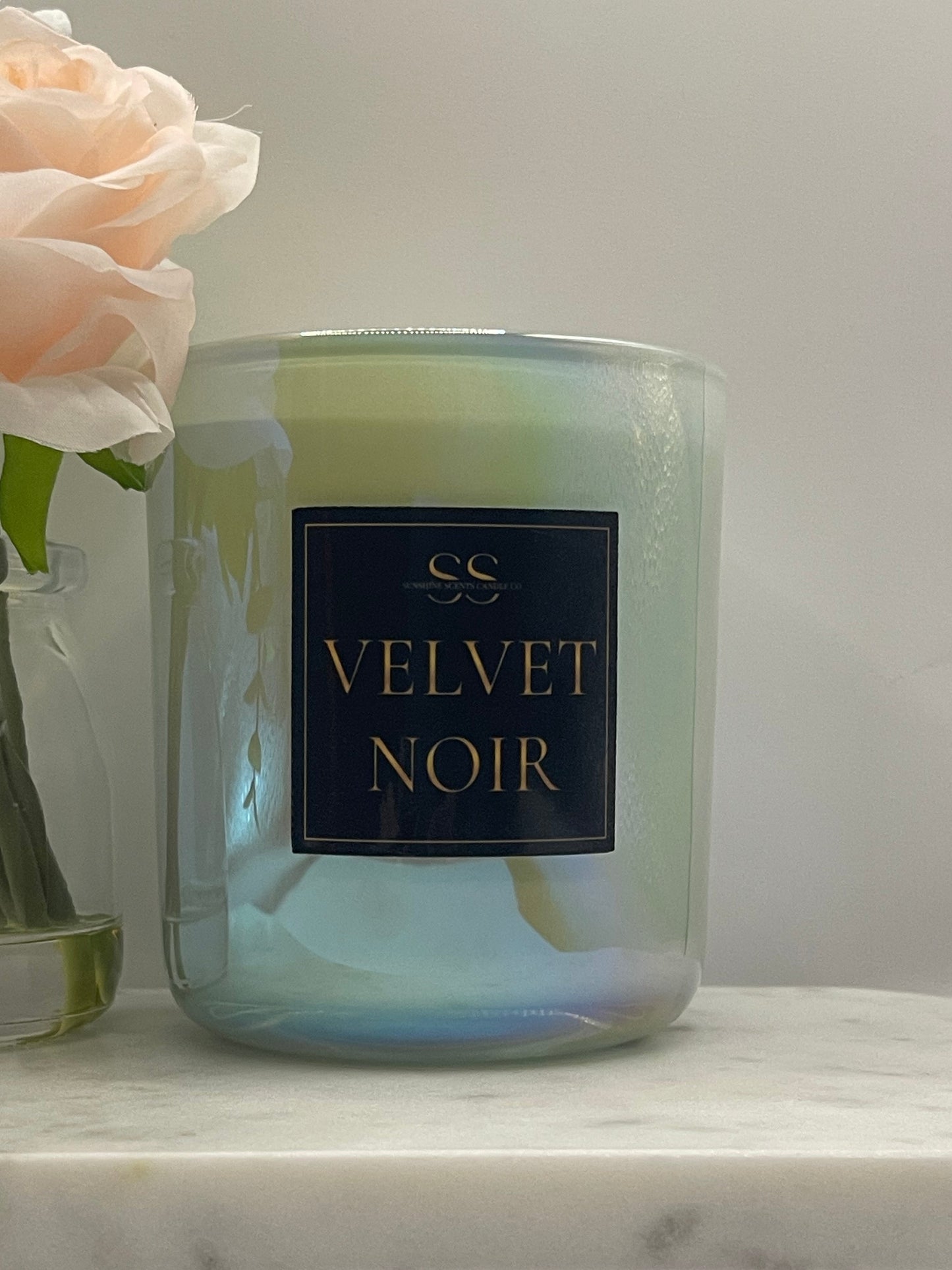 A glass jar candle with an iridescent pale green and blue finish, featuring a black label with gold lettering that reads 'VELVET NOIR'. The candle is styled on a white marble surface next to a pale pink rose.