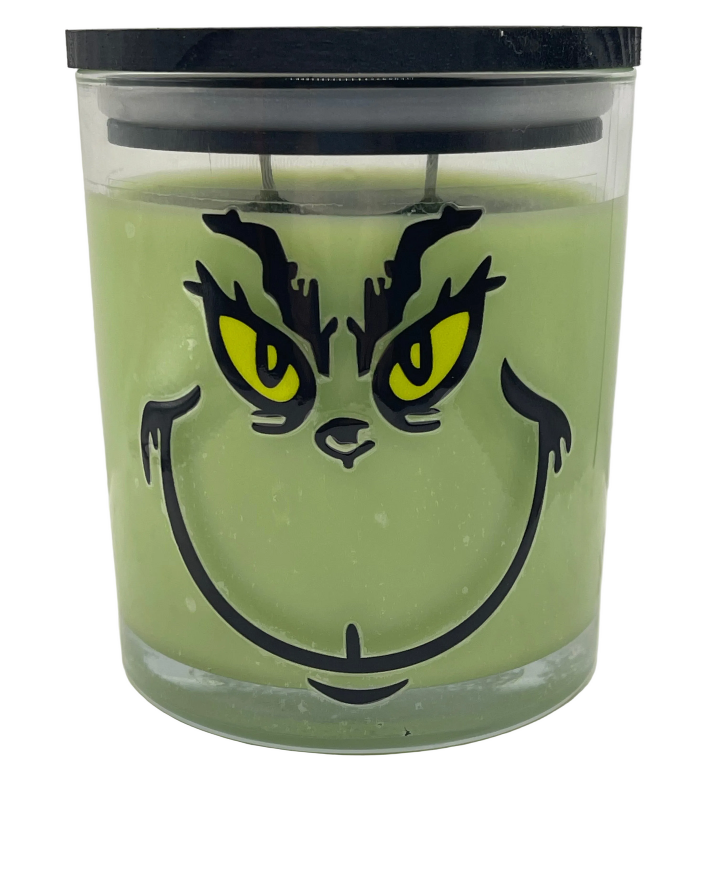 A green candle in a clear glass jar with a black lid, featuring a Grinch-style face graphic with yellow eyes printed on the front.