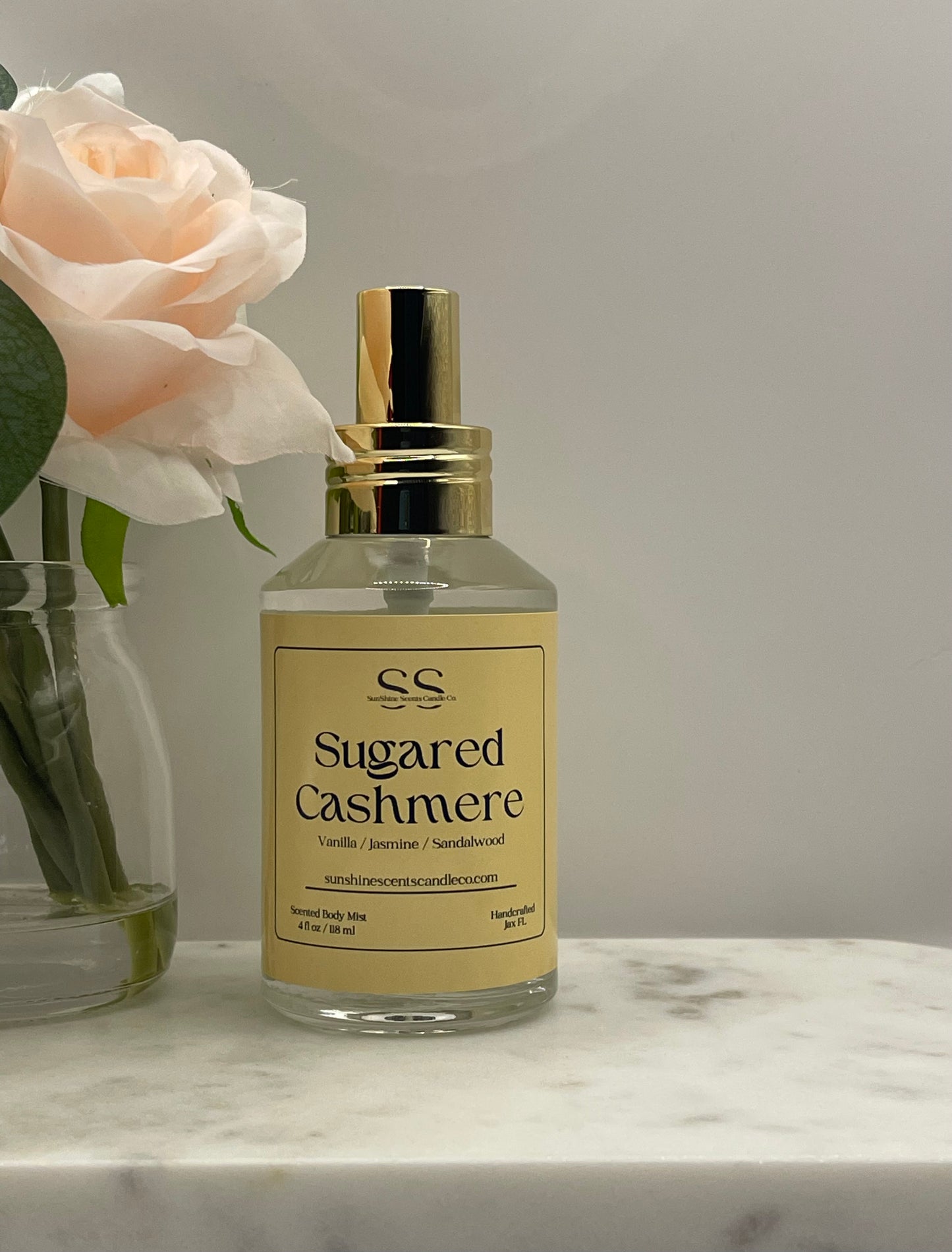 A clear glass bottle of 'Sugared Cashmere' fragrance by Sweet Southern Scents, featuring a gold spray nozzle and a beige label, set on a white marble surface next to a pale pink rose in a glass vase.