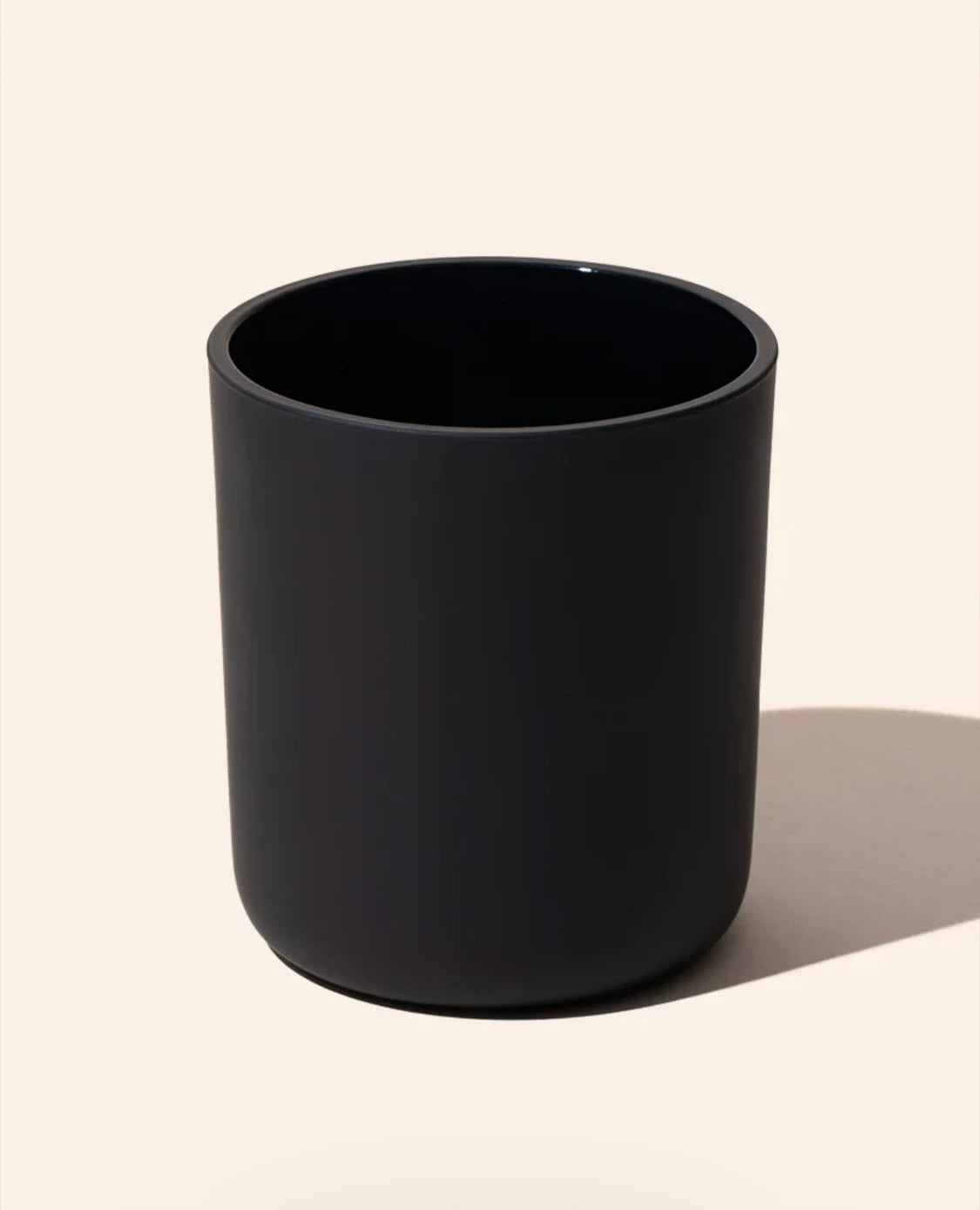 A matte black cylindrical cup or tumbler with a wide rim, shown on a warm off-white background with a soft shadow to the right.