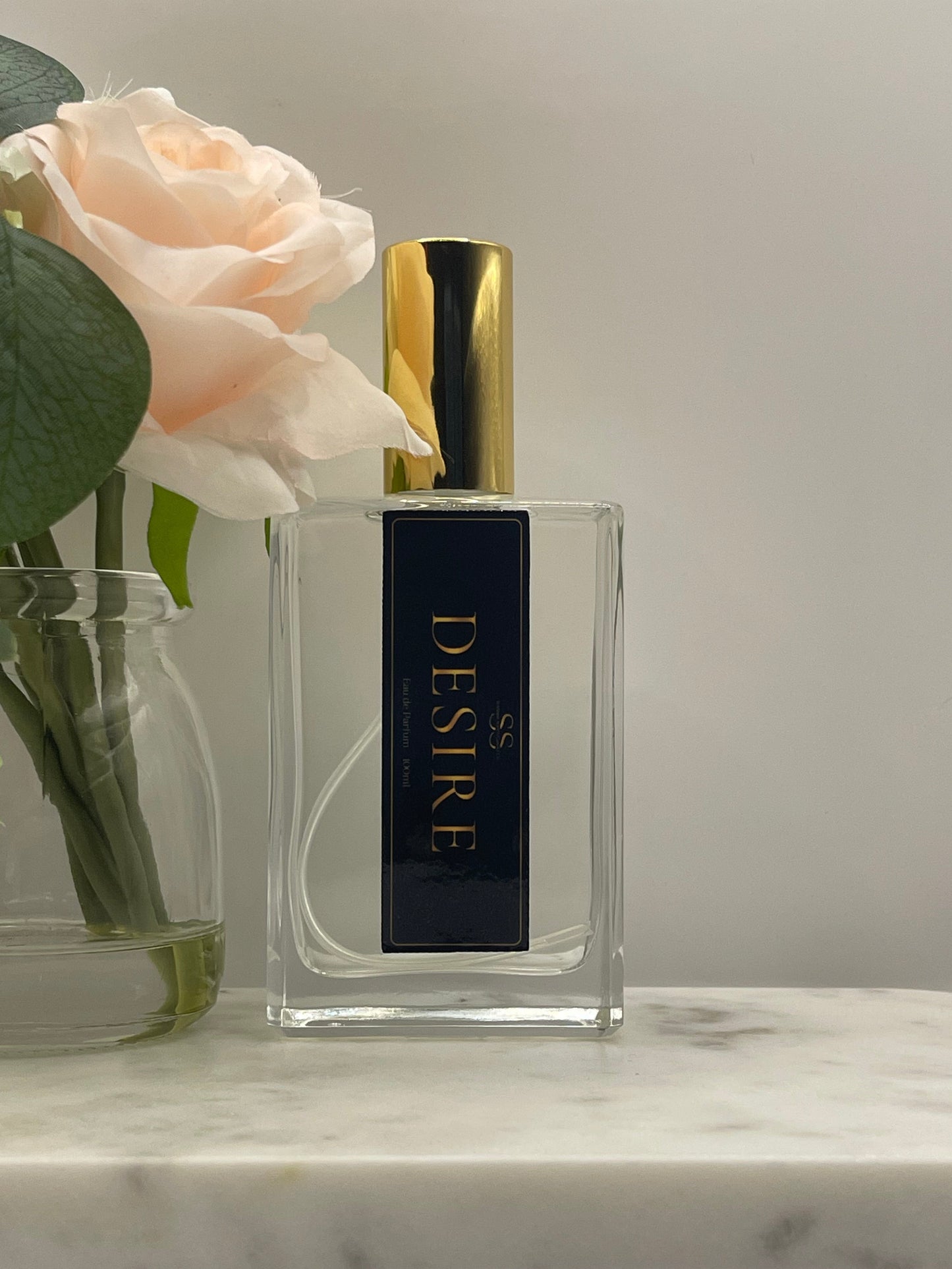 A rectangular clear glass perfume bottle with a gold cap and a dark navy label reading 'SS DESIRE' in gold, styled with pale pink roses and green leaves against a light neutral background.