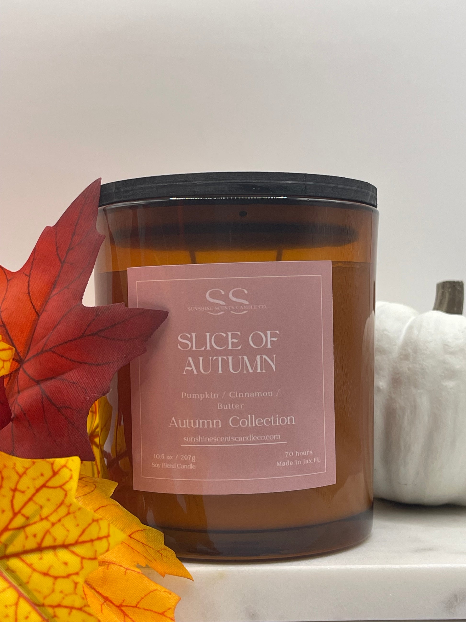 An amber glass jar candle with a dusty pink label reading 'Slice of Autumn' and 'Autumn Collection,' styled with red and yellow autumn leaves and a white decorative object in the background.