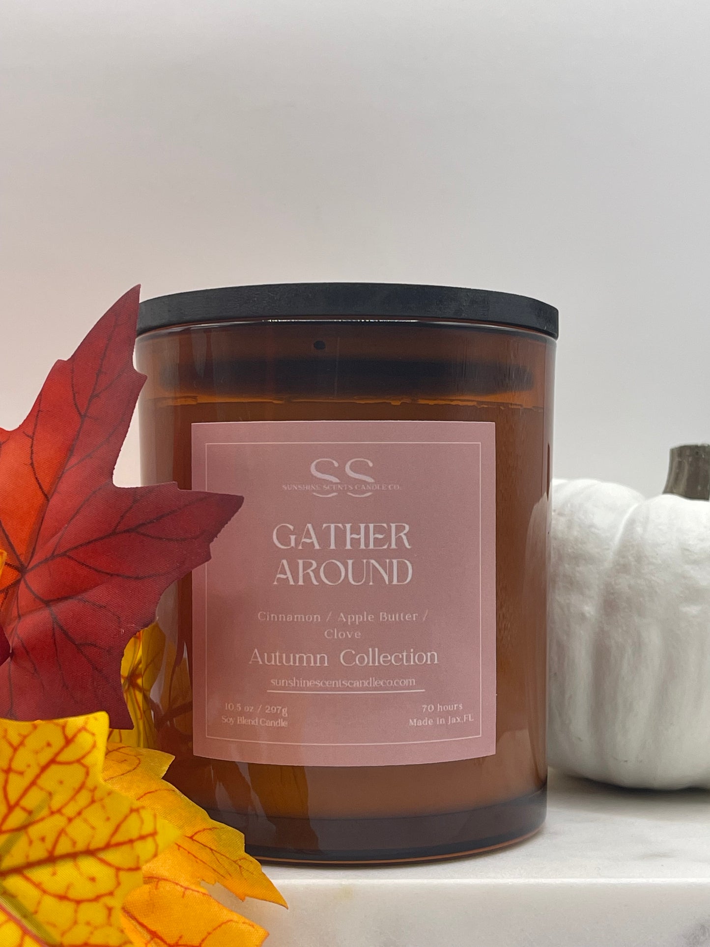 An amber glass jar candle with a black lid and a dusty pink label reading 'GATHER AROUND' and 'Autumn Collection' by SS, styled with autumn leaves and a white pumpkin on a white surface.