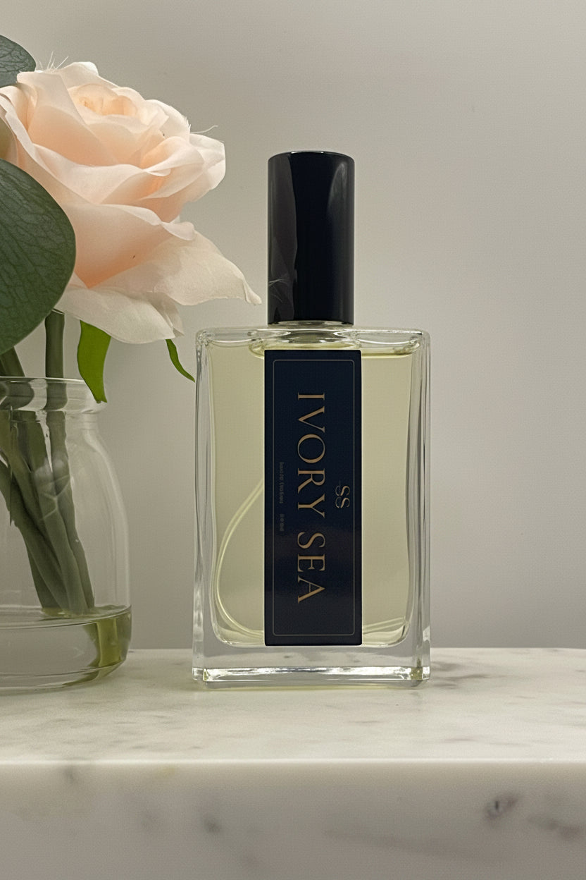 A bottle of SS Ivory Sea perfume with a black cap and a dark navy label with gold lettering, resting on a marble surface next to a pale peach rose.