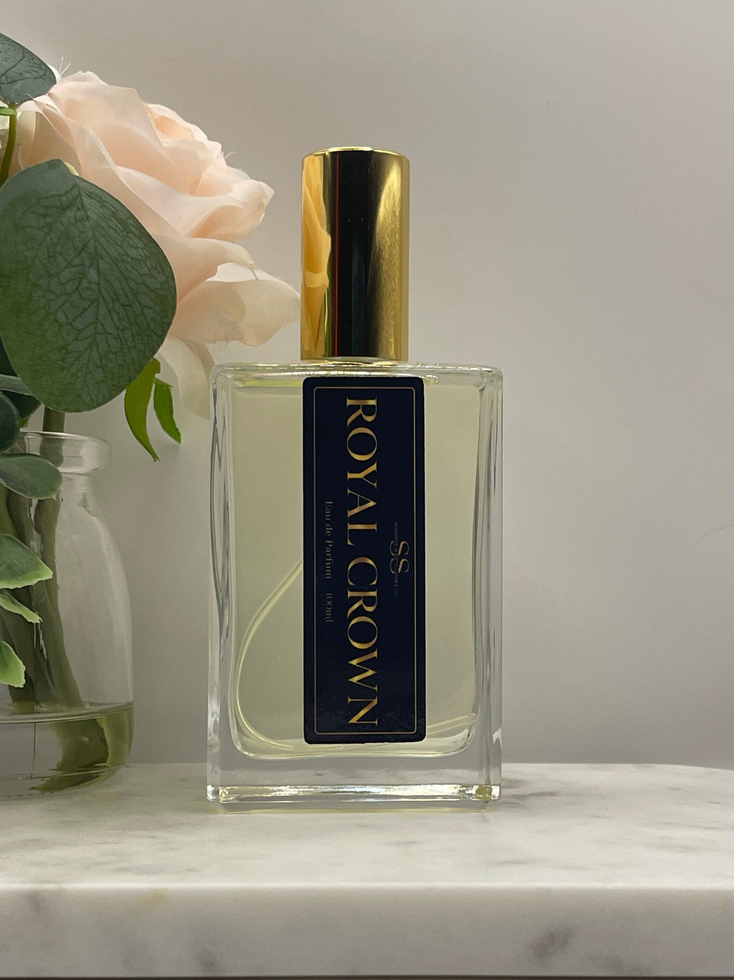 A rectangular glass bottle of Royal Crown Eau de Parfum with a gold cap and a black label, styled on a white marble surface next to a pale pink rose and green leaves.