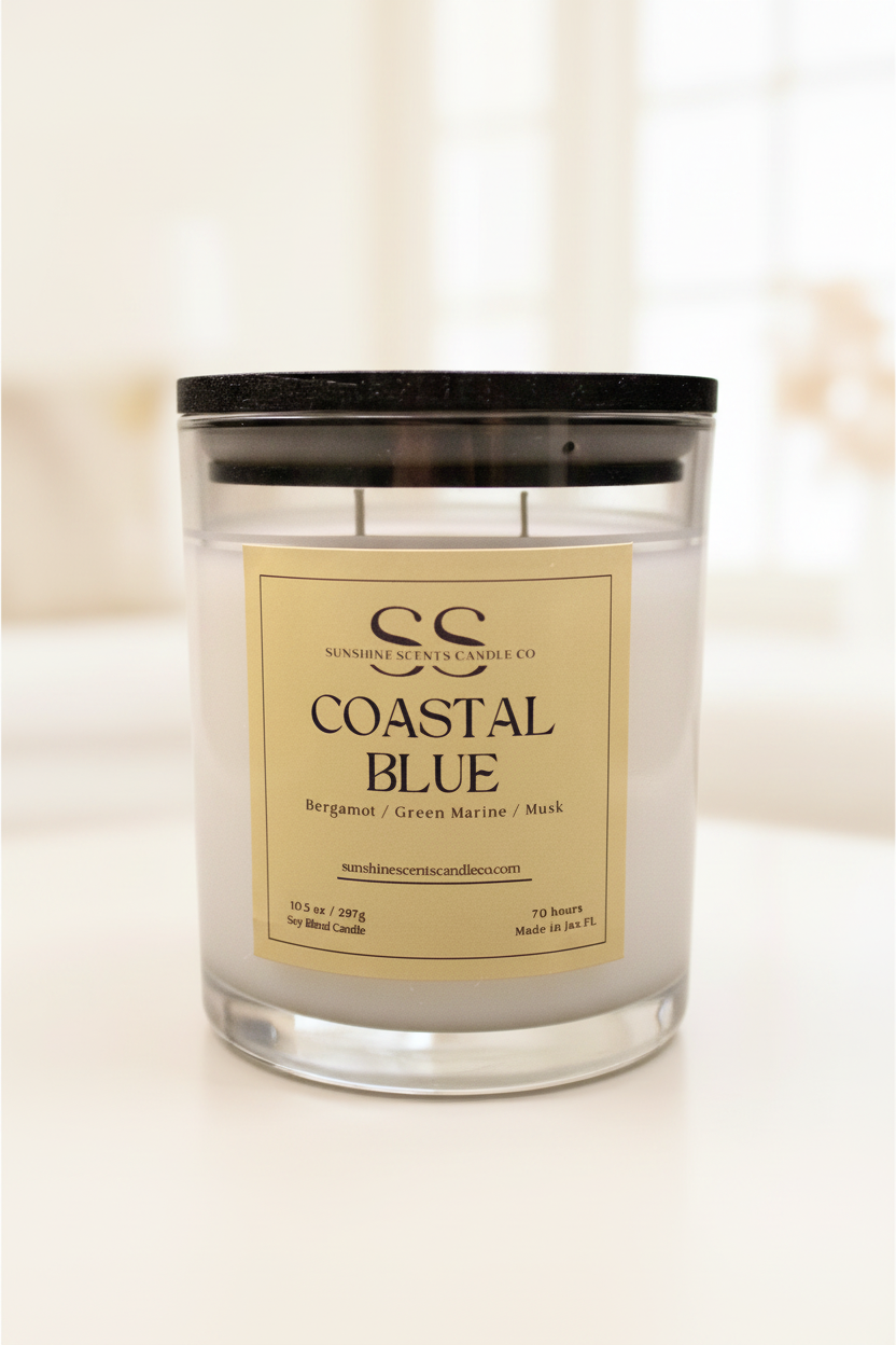 A glass jar candle with a black lid and a cream-colored label reading 'COASTAL BLUE' by Sunshine Scents Candle Co, set against a bright, softly blurred indoor background.