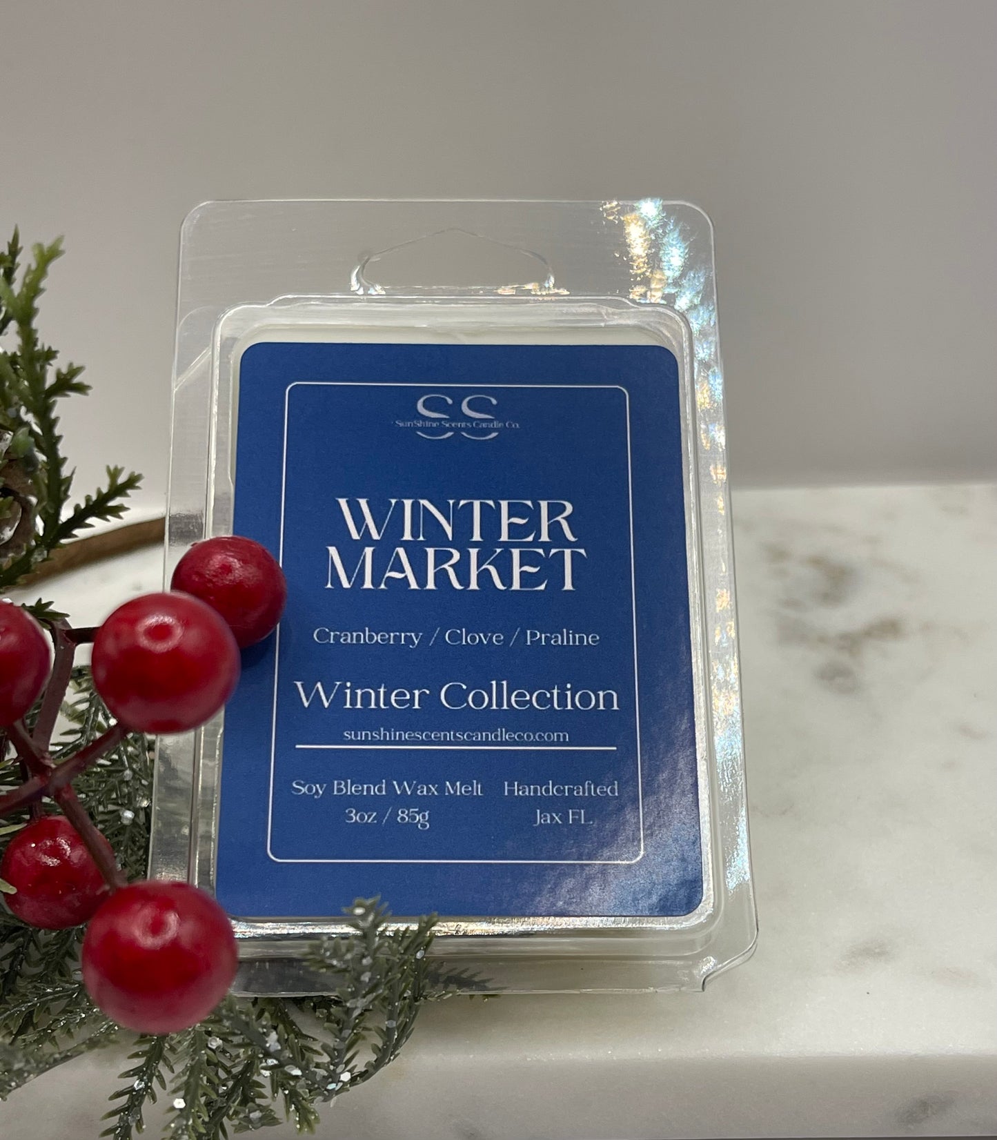 A packaged wax melt product labeled 'Winter Market' from Sunshine Scents, shown in a clear plastic clamshell package with a blue front label. The package is photographed on a light marble surface with holiday greenery and red berries beside it.