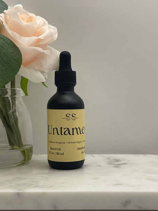 A dark glass dropper bottle with a beige label reading 'Untamed' by 'ec', resting on a white marble surface next to a pale peach rose in a glass vase.