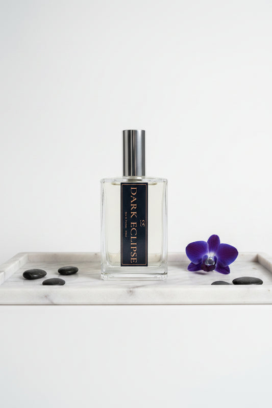 "Dark Eclipse Eau de Parfum glass bottle with silver cap and black label, styled on a marble tray with black stones and a purple orchid on a white background — luxury perfume by SunShine Scents Candle Co."