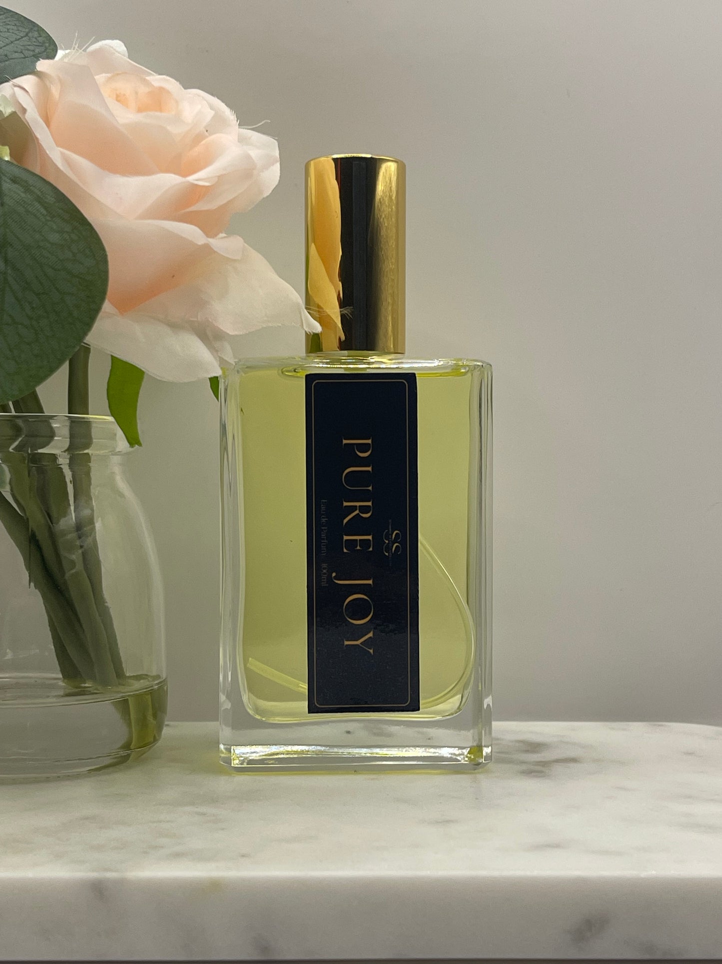 A rectangular glass perfume bottle with pale yellow liquid, a shiny gold cap, and a dark navy label reading 'PURE JOY' in gold lettering. The bottle is styled with a pale peach rose and green leaves against a light gray background and a white marble surface.