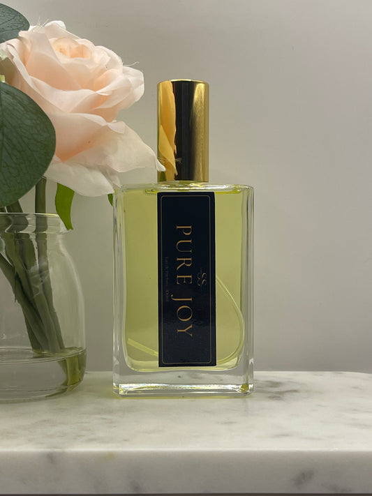 A rectangular glass perfume bottle with pale yellow liquid, a shiny gold cap, and a dark navy label reading 'PURE JOY' in gold lettering. The bottle is styled with a pale peach rose and green leaves against a light gray background and a white marble surface.