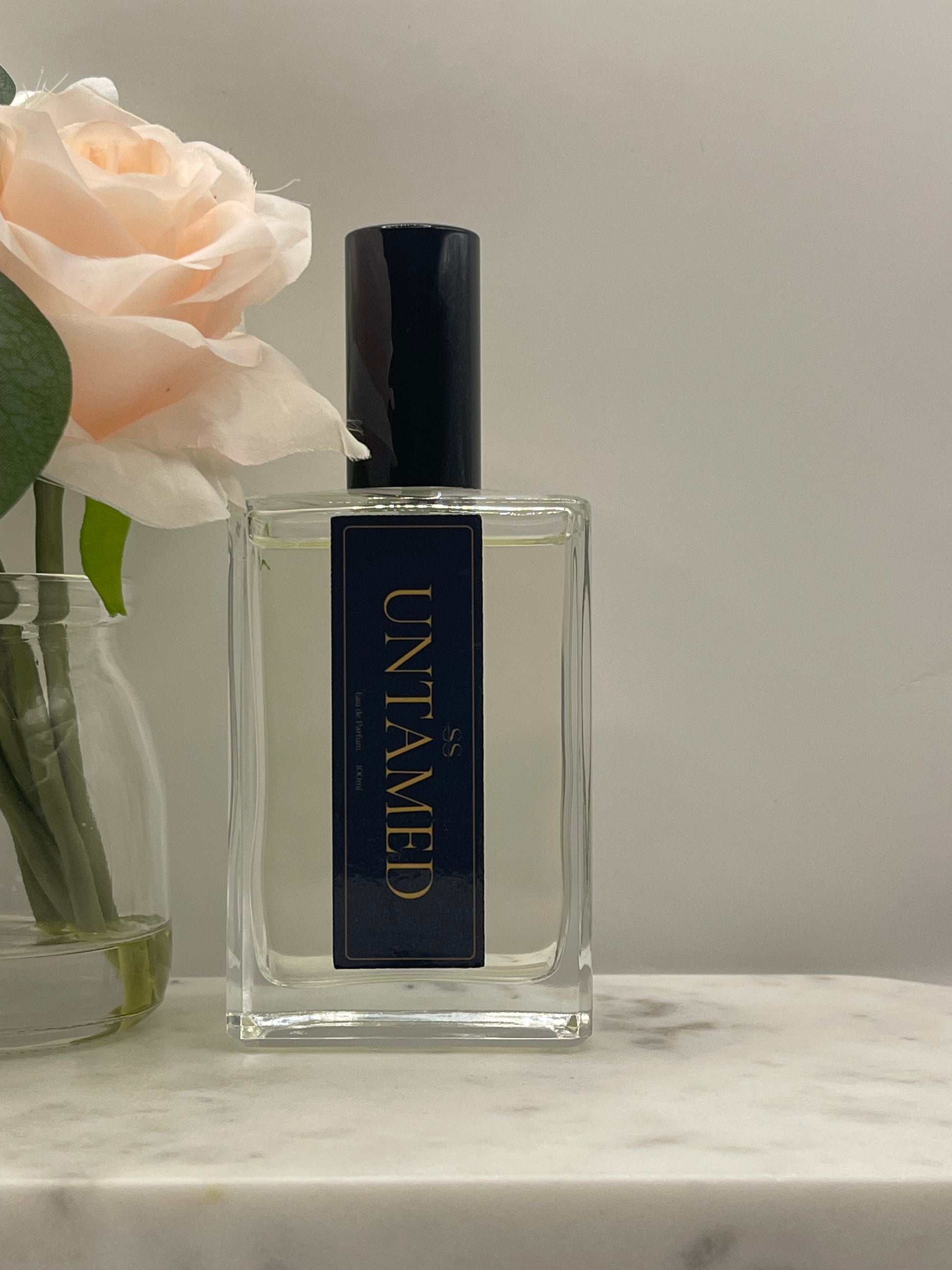 A clear rectangular glass perfume bottle with a black cap and a black label featuring gold lettering that reads 'UNTAMED'. The bottle is styled on a white marble surface next to a pale peach rose.
