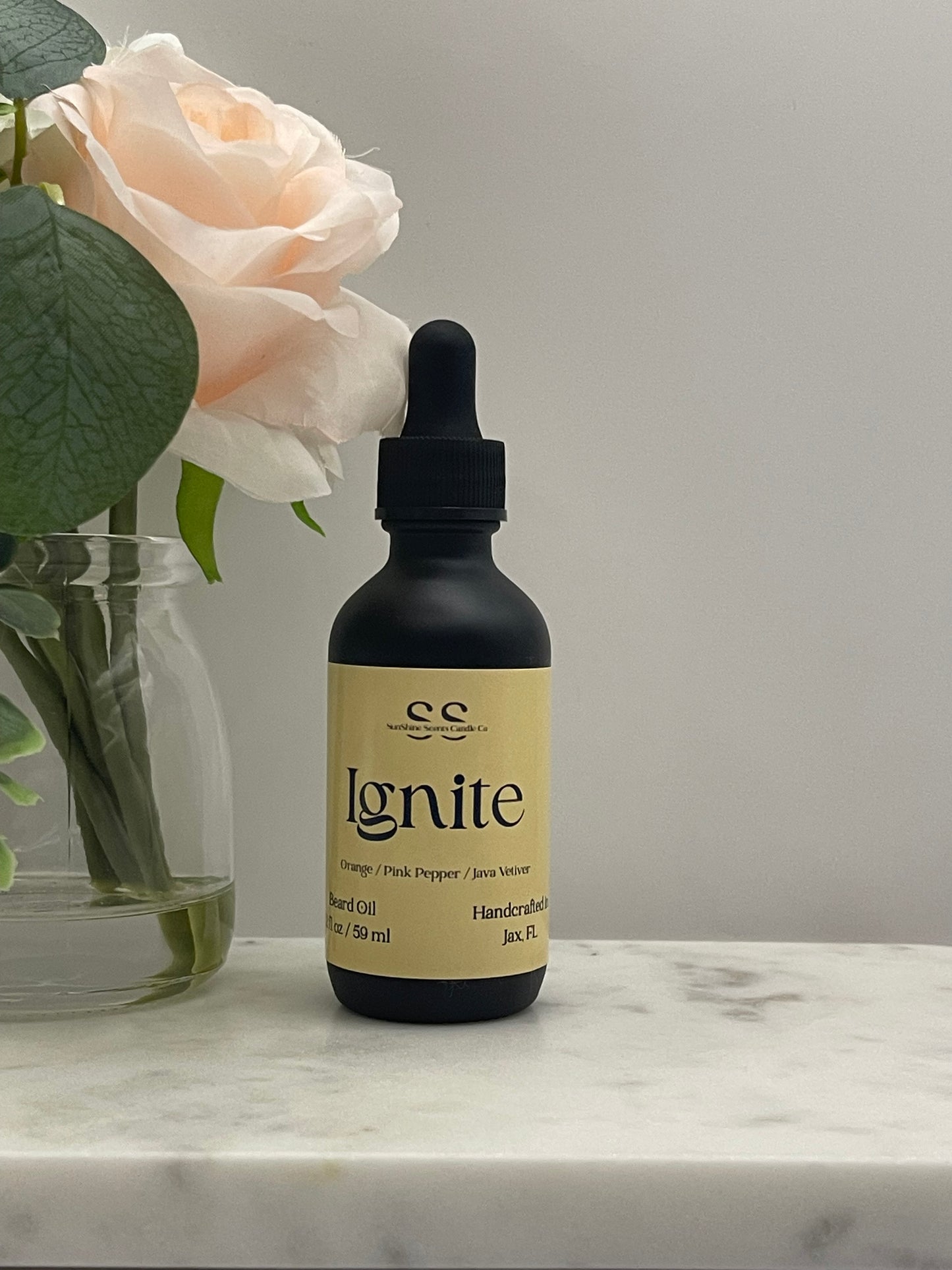 A small black dropper bottle with a beige label reading 'ignite' and 'handcrafted' by 'es', resting on a white marble surface next to a pale pink rose in a glass vase.