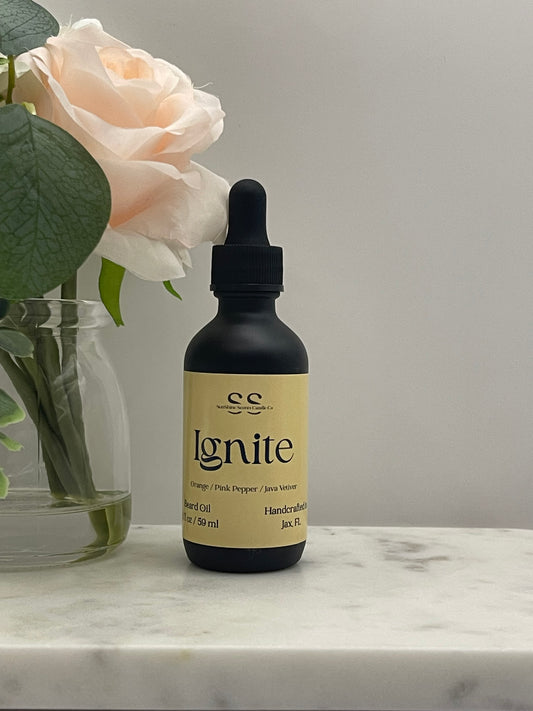 A small black dropper bottle with a beige label reading 'ignite' and 'handcrafted' by 'es', resting on a white marble surface next to a pale pink rose in a glass vase.