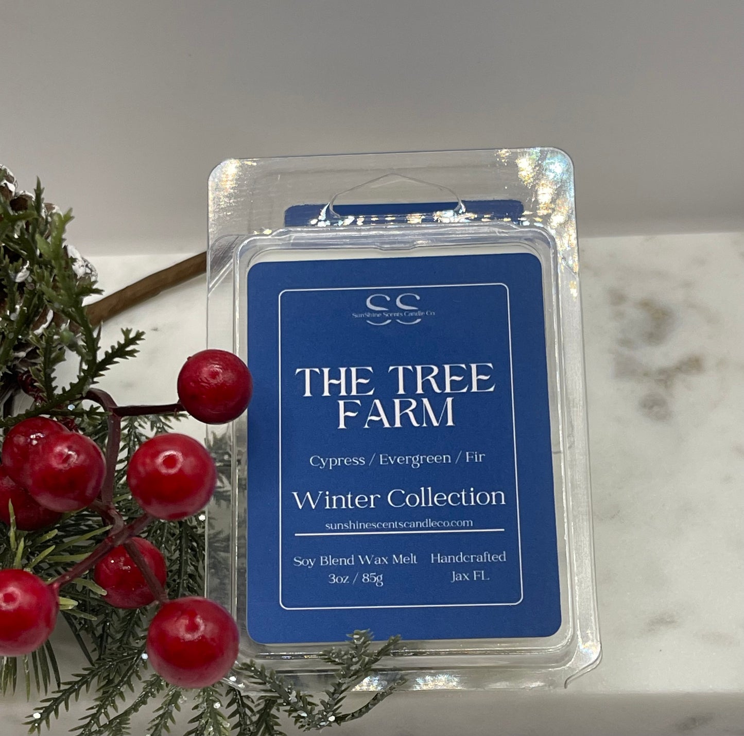 A packaged wax melt product labeled 'THE TREE FARM Winter Collection' sits on a light marble-like surface with evergreen branches and red berries beside it. The product is in a clear plastic clamshell with a blue front label.