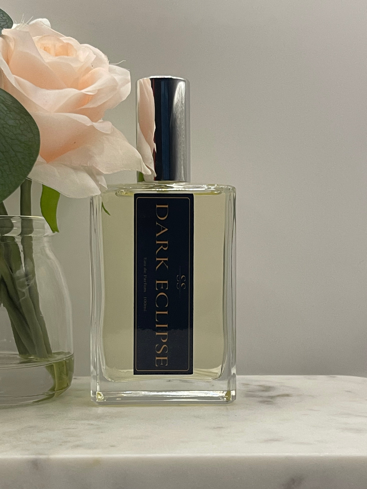 A rectangular glass perfume bottle labeled 'DARK ECLIPSE' with a dark navy label and silver cap, placed on a marble surface beside a pale pink rose in a glass vase against a soft gray background.