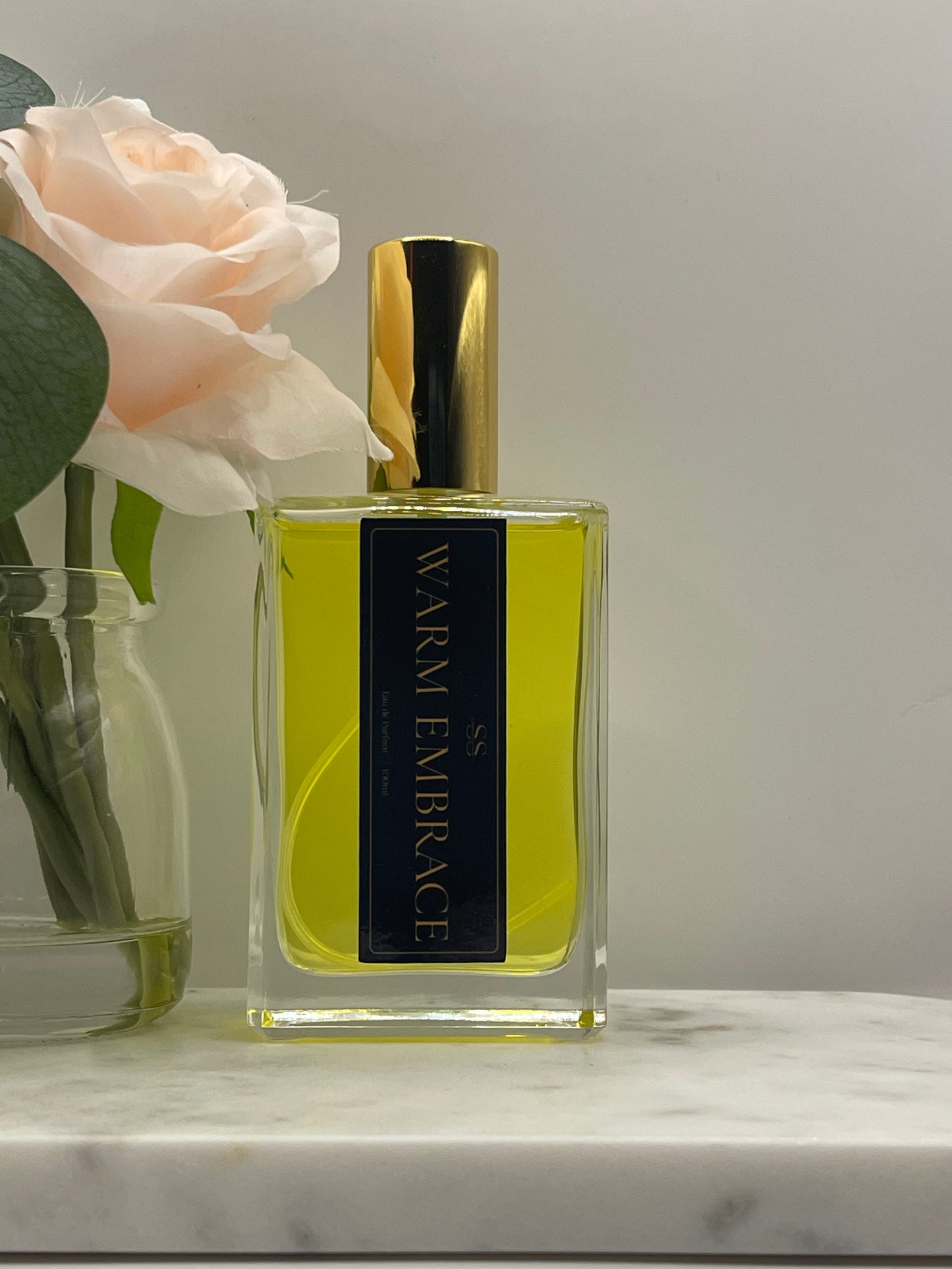 A rectangular glass perfume bottle with a gold cap and a black label reading 'WARM EMBRACE' in gold lettering, filled with yellow-green liquid. The bottle is styled with a pale peach rose and green leaves against a light gray background.