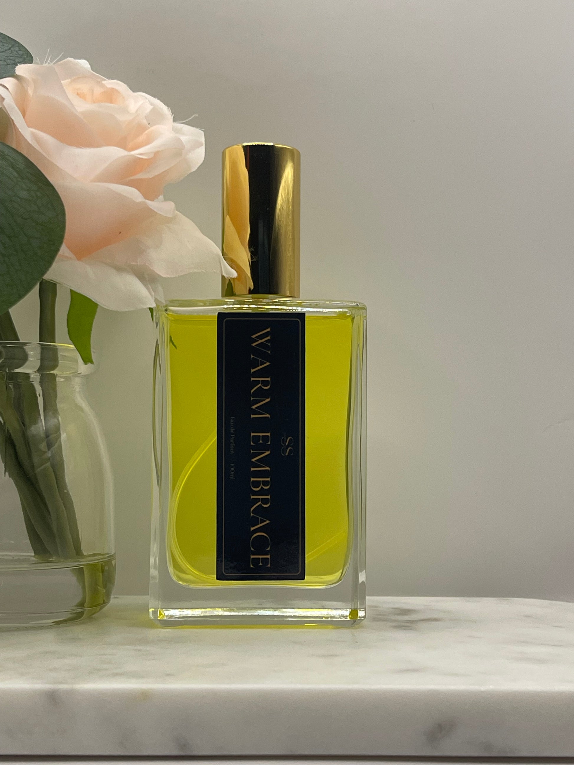 A rectangular glass perfume bottle with a gold cap and a black label reading 'WARM EMBRACE' in gold lettering, filled with yellow-green liquid. The bottle is styled with a pale peach rose and green leaves against a light gray background.