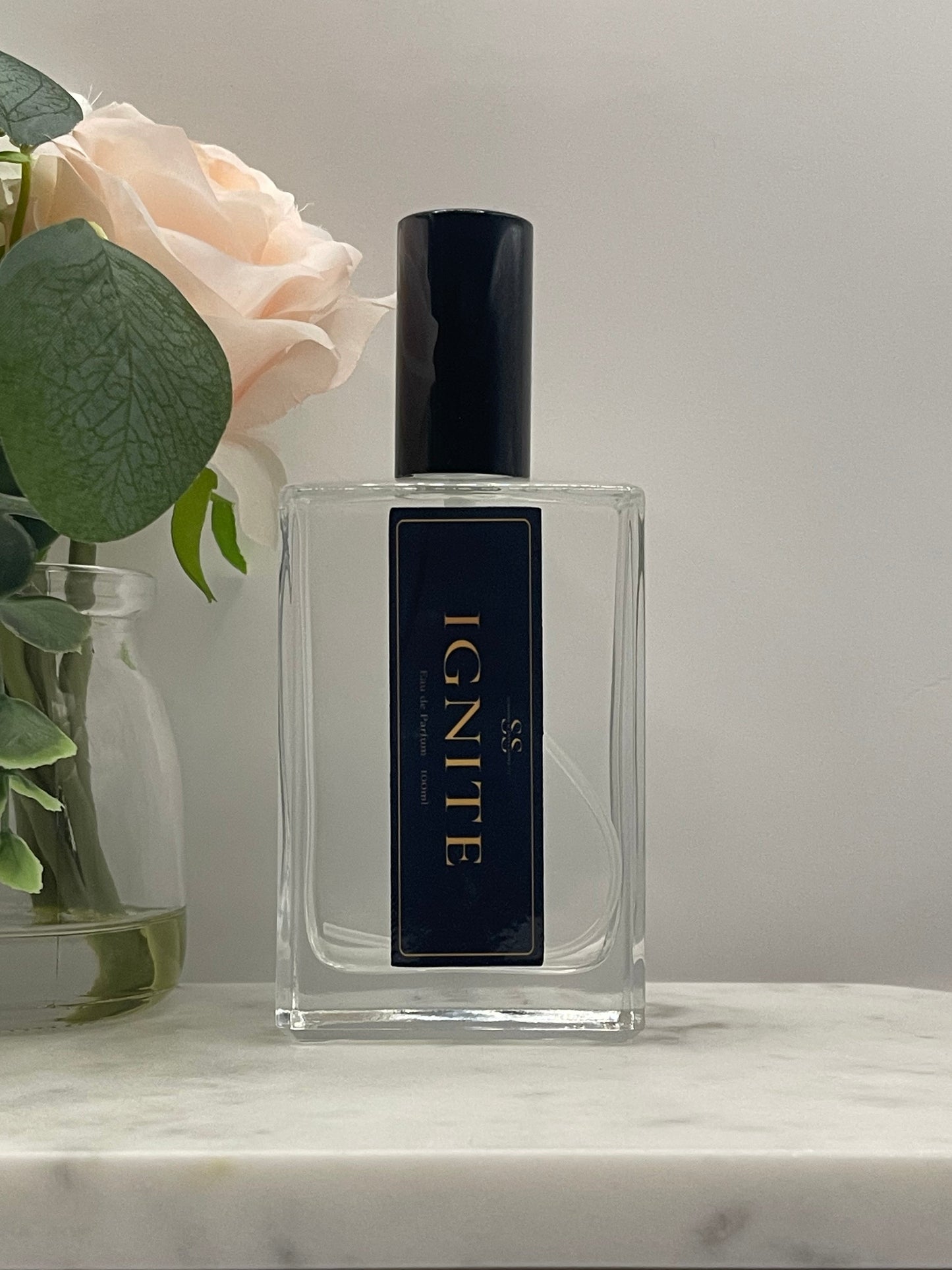 A clear rectangular perfume bottle with a black cap and a black label reading IGNITE in gold lettering, styled with a pale pink rose and green leaves against a light gray background.