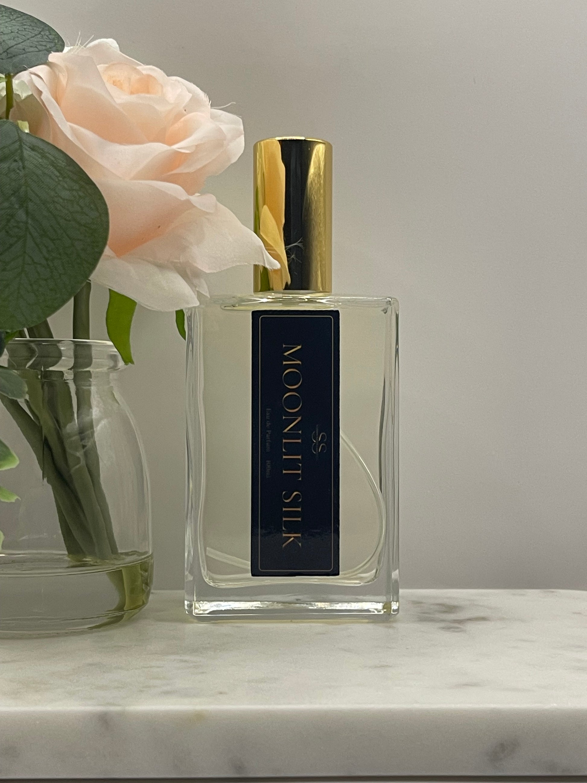 A rectangular glass perfume bottle labeled 'MOONLIT SILK' with a gold cap, shown beside a pale blush rose and green leaves against a light gray background.