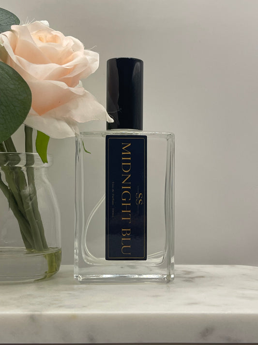 A clear rectangular perfume bottle labeled 'SS MIDNIGHT BLU' in gold lettering on a dark navy label, with a black cap. The bottle is styled with a pale peach rose and green leaves against a light gray background and a white marble surface.