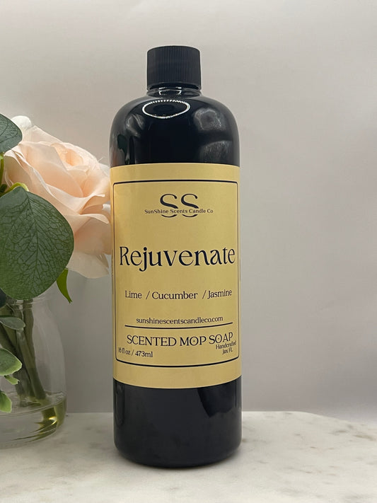 A bottle of SS Scents Rejuvenate Scented Mop Soap, featuring a black bottle and a beige label, styled with a pale peach rose and green leaves on a light marble surface.