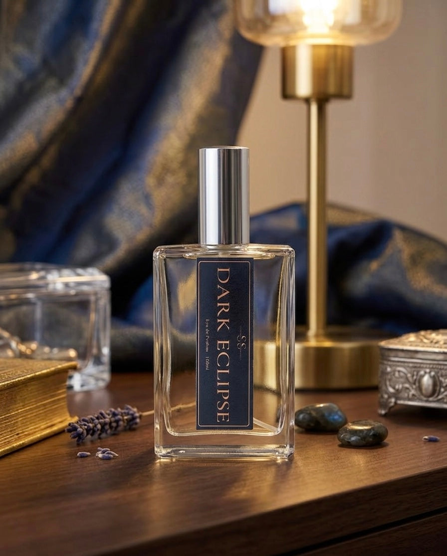 "Dark Eclipse Eau de Parfum 100ml bottle on a wooden vanity surrounded by lavender sprigs, dark stones, and a glowing brass lamp with deep blue silk fabric — small-batch luxury fragrance by SunShine Scents Candle Co."