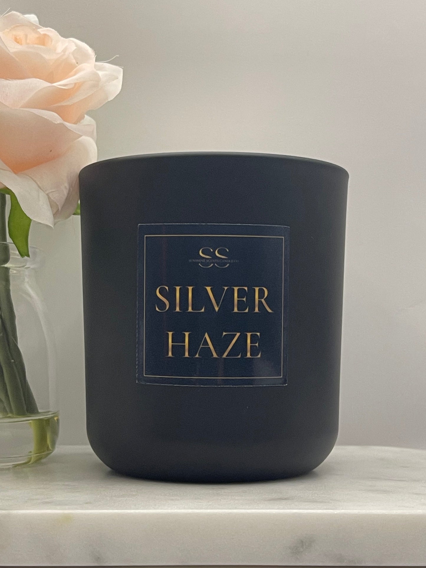 A matte black candle jar with a gold and navy label reading 'SILVER HAZE' and a small monogram logo, photographed against a light neutral background with a pale rose and glass vase at the lower left.