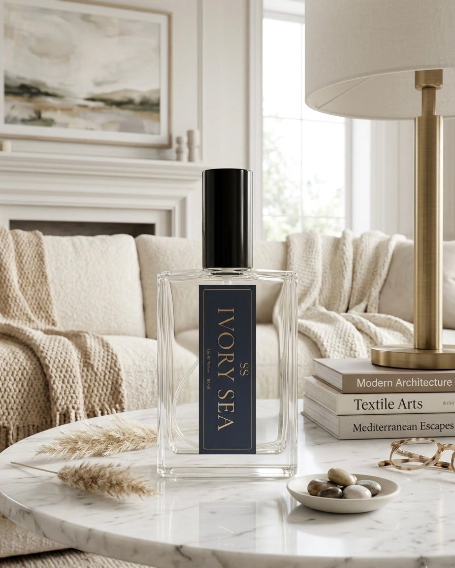 "Ivory Sea Eau de Parfum 100ml bottle by SunShine Scents Candle Co. on a marble coffee table with dried pampas grass, smooth stones, and stacked books including Mediterranean Escapes, in a bright coastal living room."