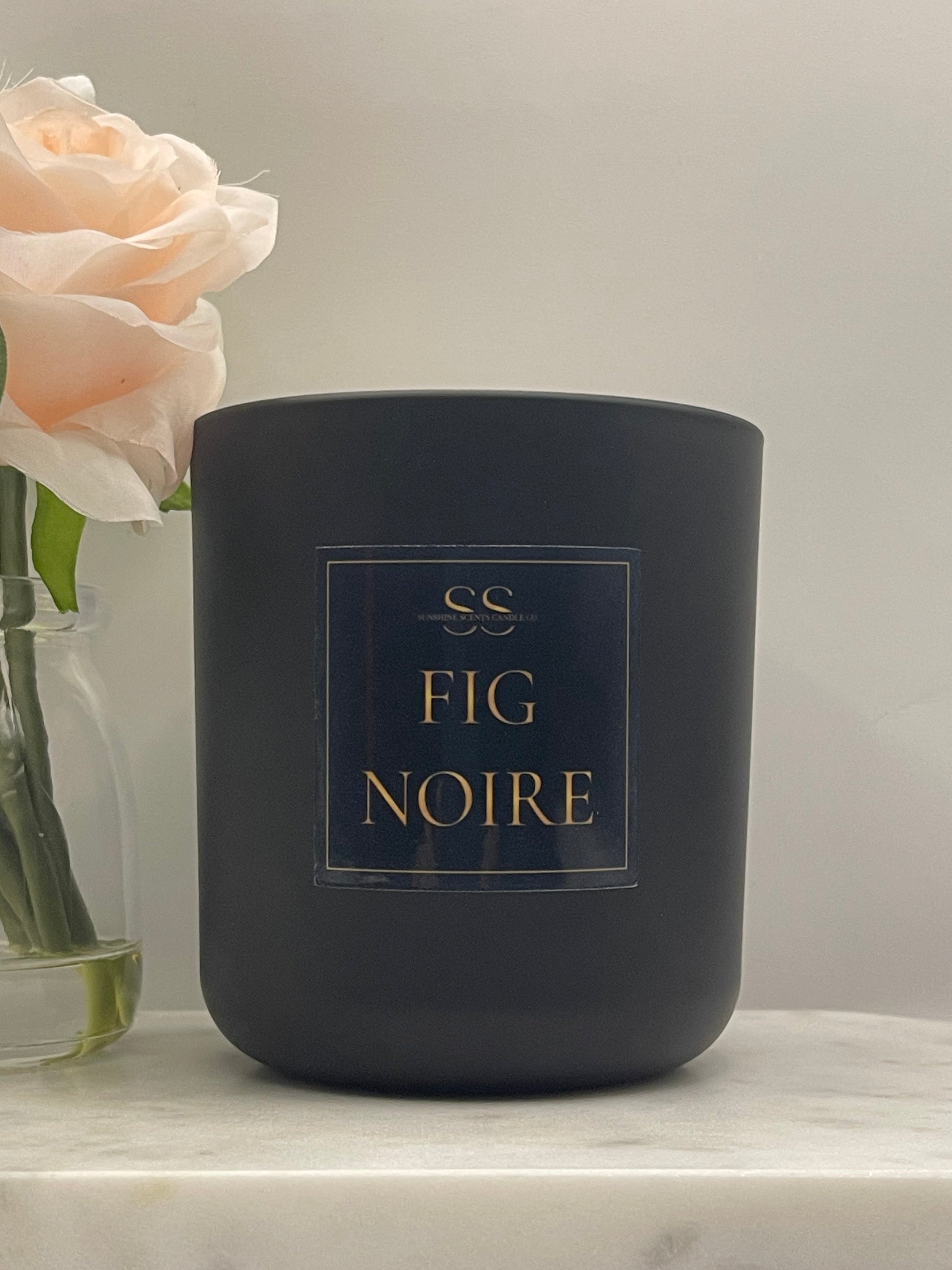 A matte black candle jar with gold lettering that reads 'SS Fig Noire' on a marble surface next to a pale pink rose in a glass vase.