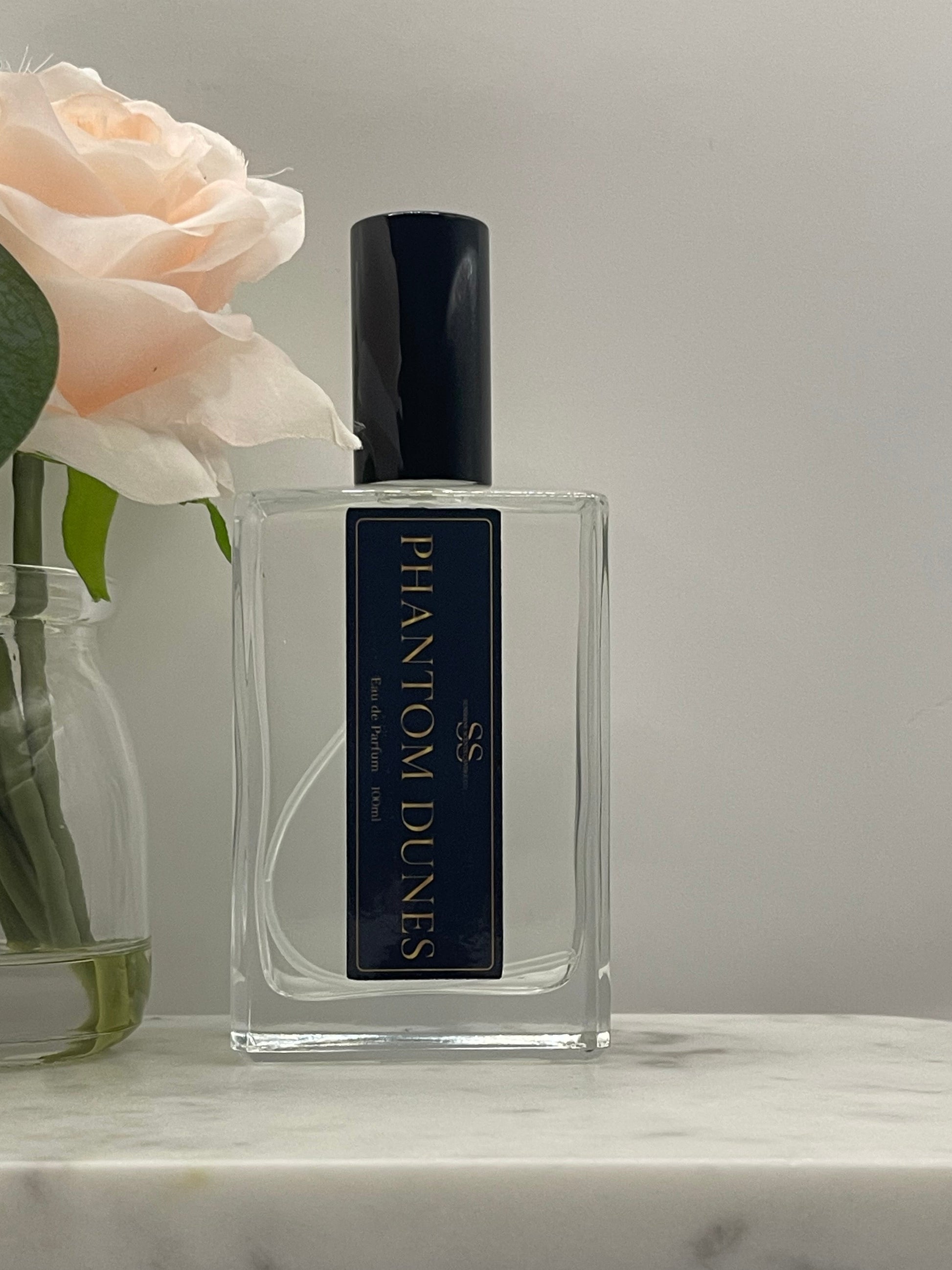 A clear rectangular glass perfume bottle with a black cap and a black label reading PHANTOM DUNES, styled with a pale peach rose in the foreground against a light gray background.