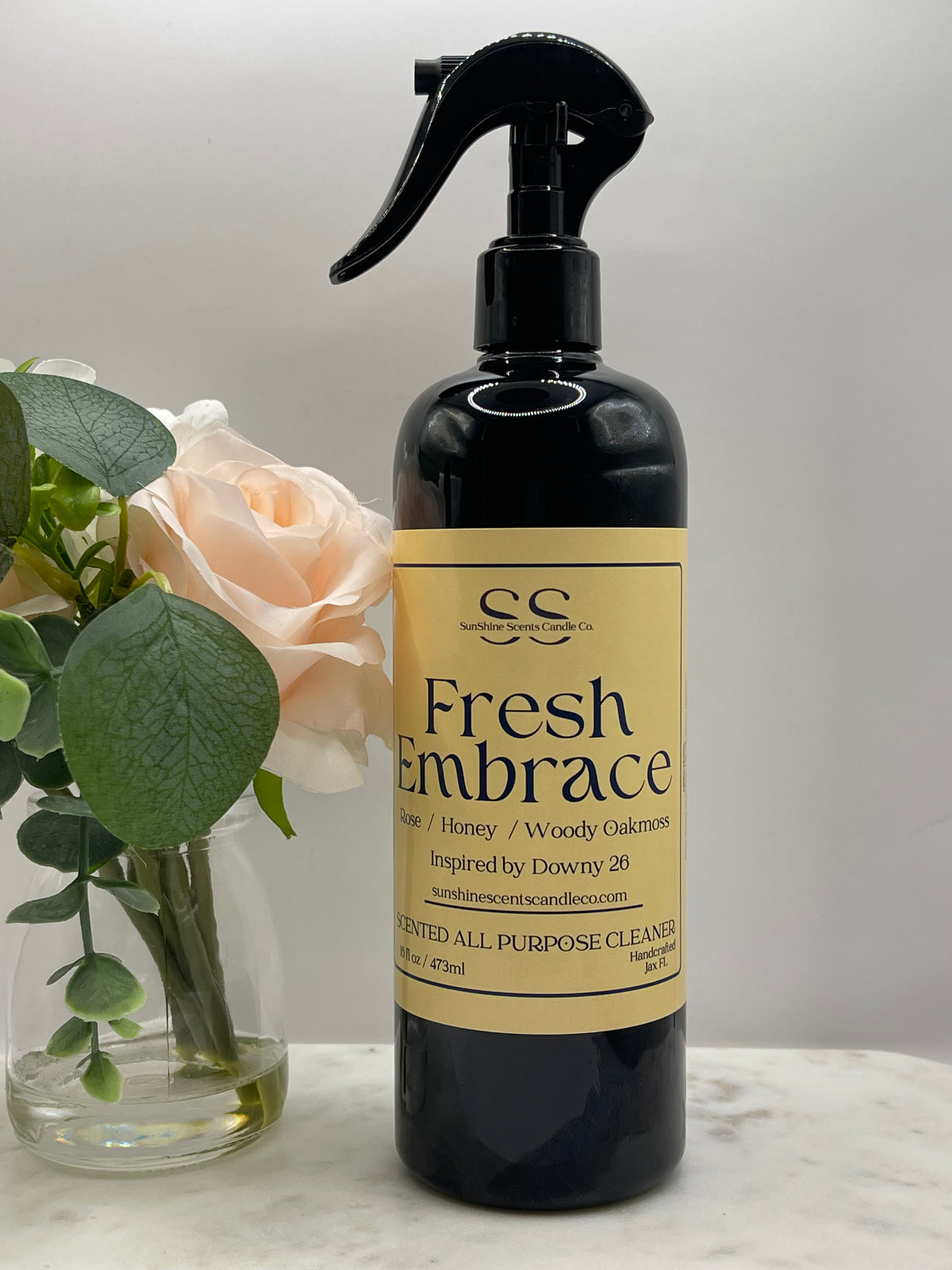 “SunShine Scents Candle Co. Fresh Embrace All Purpose Cleaner 16oz spray bottle with a yellow label beside a vase of pale roses.”