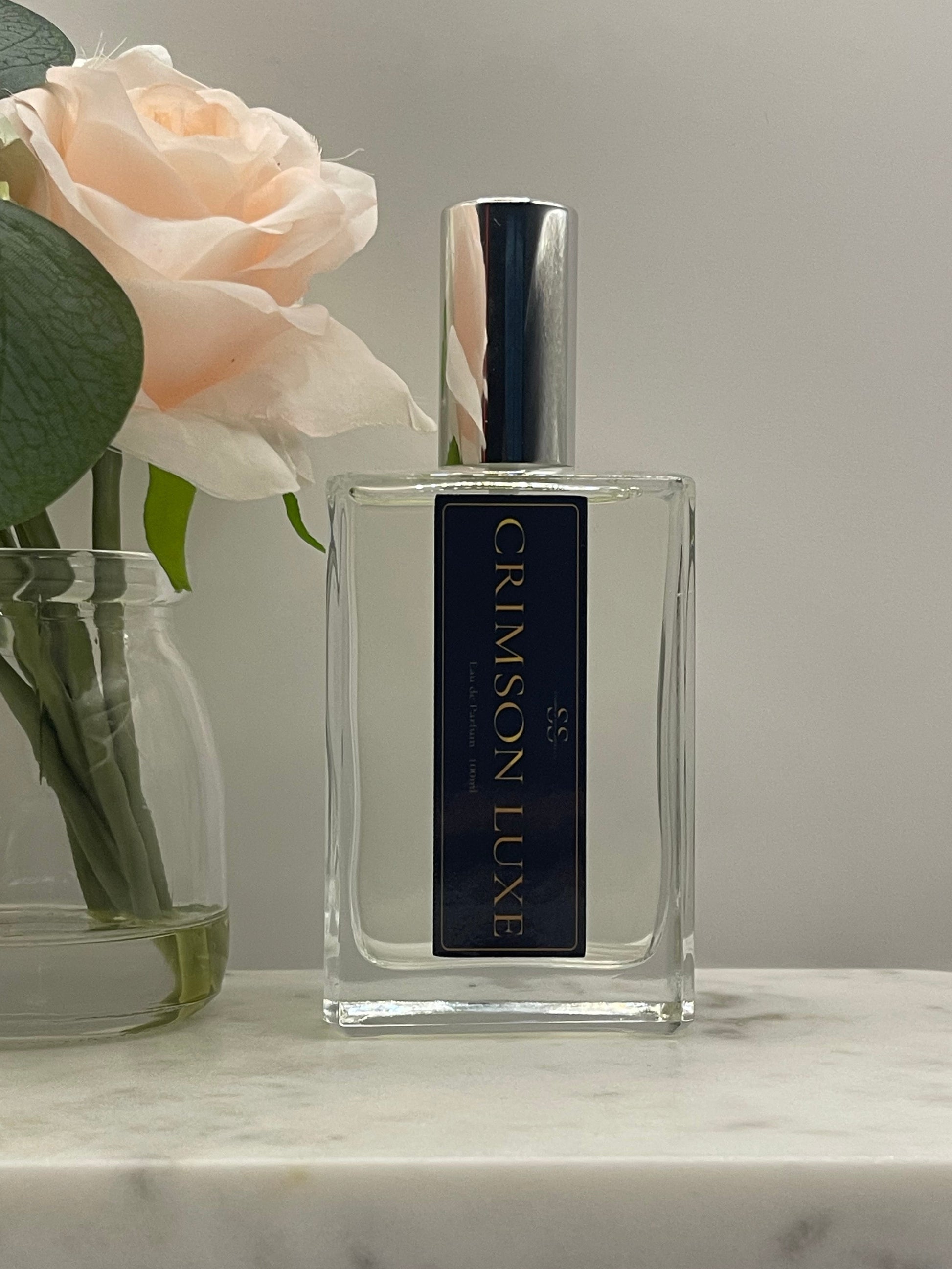 A clear rectangular perfume bottle labeled 'SC CRIMSON LUXE' in gold lettering on a black label, with a reflective silver cap. The bottle is styled with a pale peach rose and green leaves against a light gray background and a white marble surface.