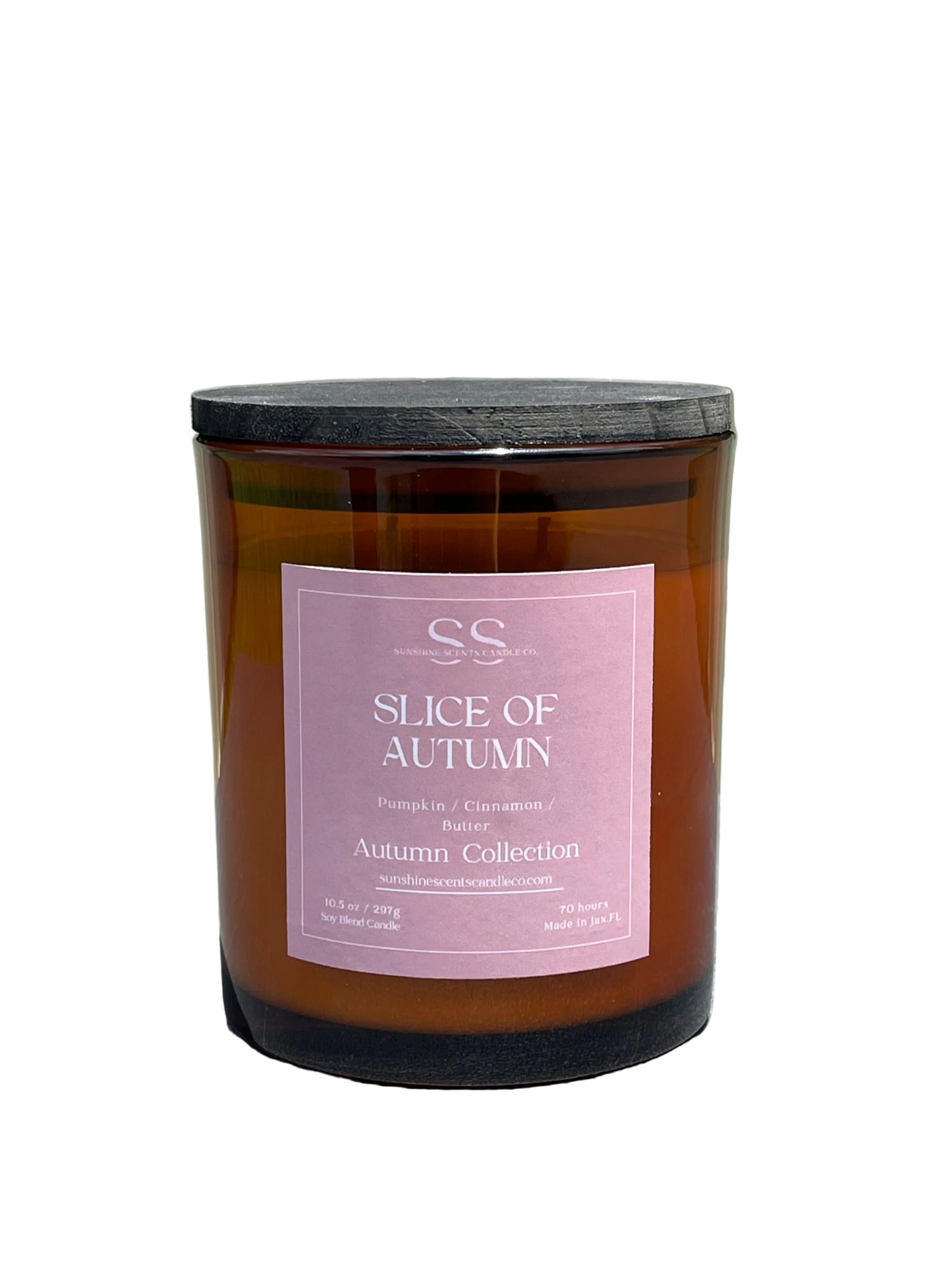 An amber glass jar candle with a black lid and a dusty pink label reading 'SS Slice of Autumn Autumn Collection,' photographed outdoors on a gray surface with a light fence and greenery blurred in the background.