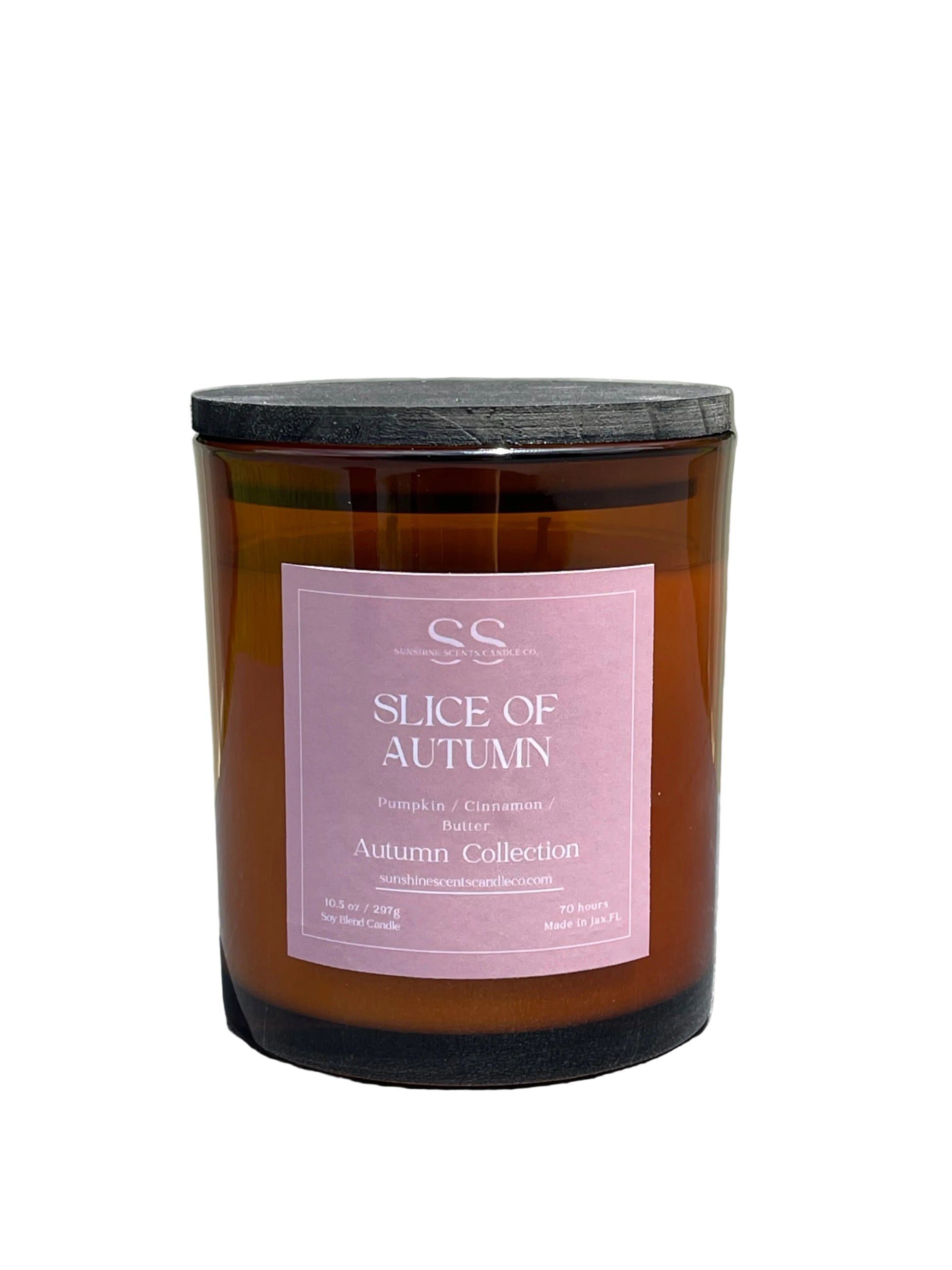 An amber glass jar candle with a black lid and a dusty pink label reading 'SS Slice of Autumn Autumn Collection,' photographed outdoors on a gray surface with a light fence and greenery blurred in the background.
