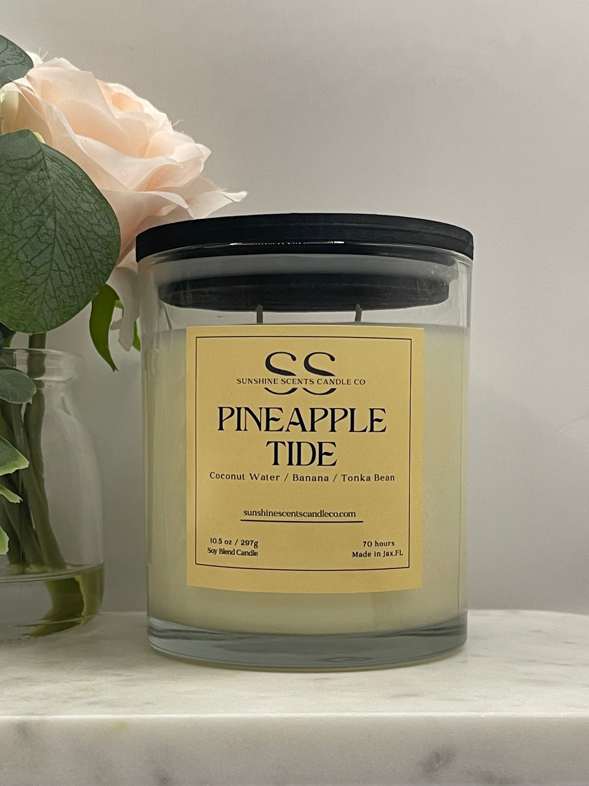 A glass jar candle with a black lid and a cream-colored label reading 'GC PINEAPPLE TIDE' and 'Coconut Water / Banana / Pineapple Scent.' The candle is photographed on a light marble-like surface with a pale flower and green leaves beside it.