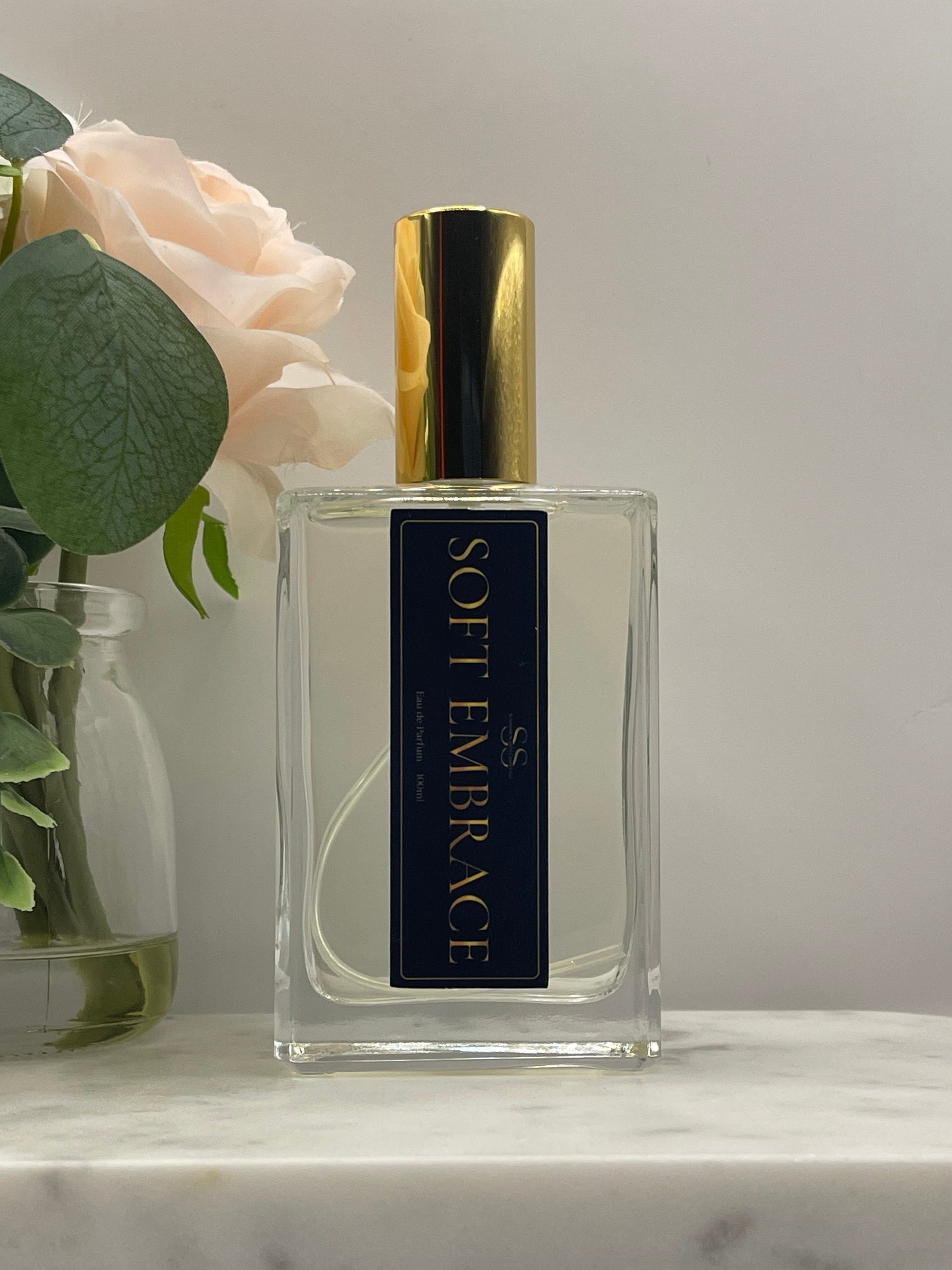 A rectangular glass perfume bottle labeled 'SOFT EMBRACE' with a gold cap, displayed against a light background with a pale pink rose and green leaves in the foreground.