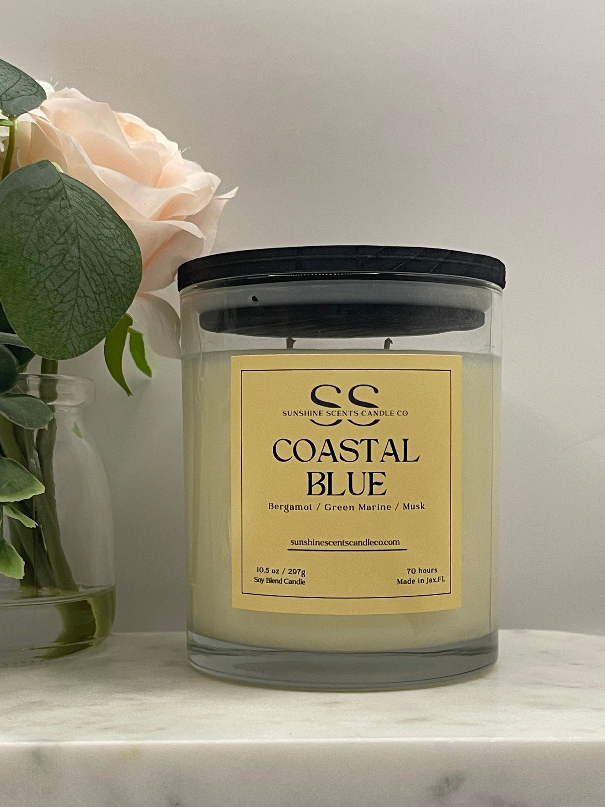 A candle in a clear glass jar with a black lid and a cream-colored label reading 'COASTAL BLUE' by Southern Tide Candle Co., styled with pale pink roses and green leaves on a light marble surface.
