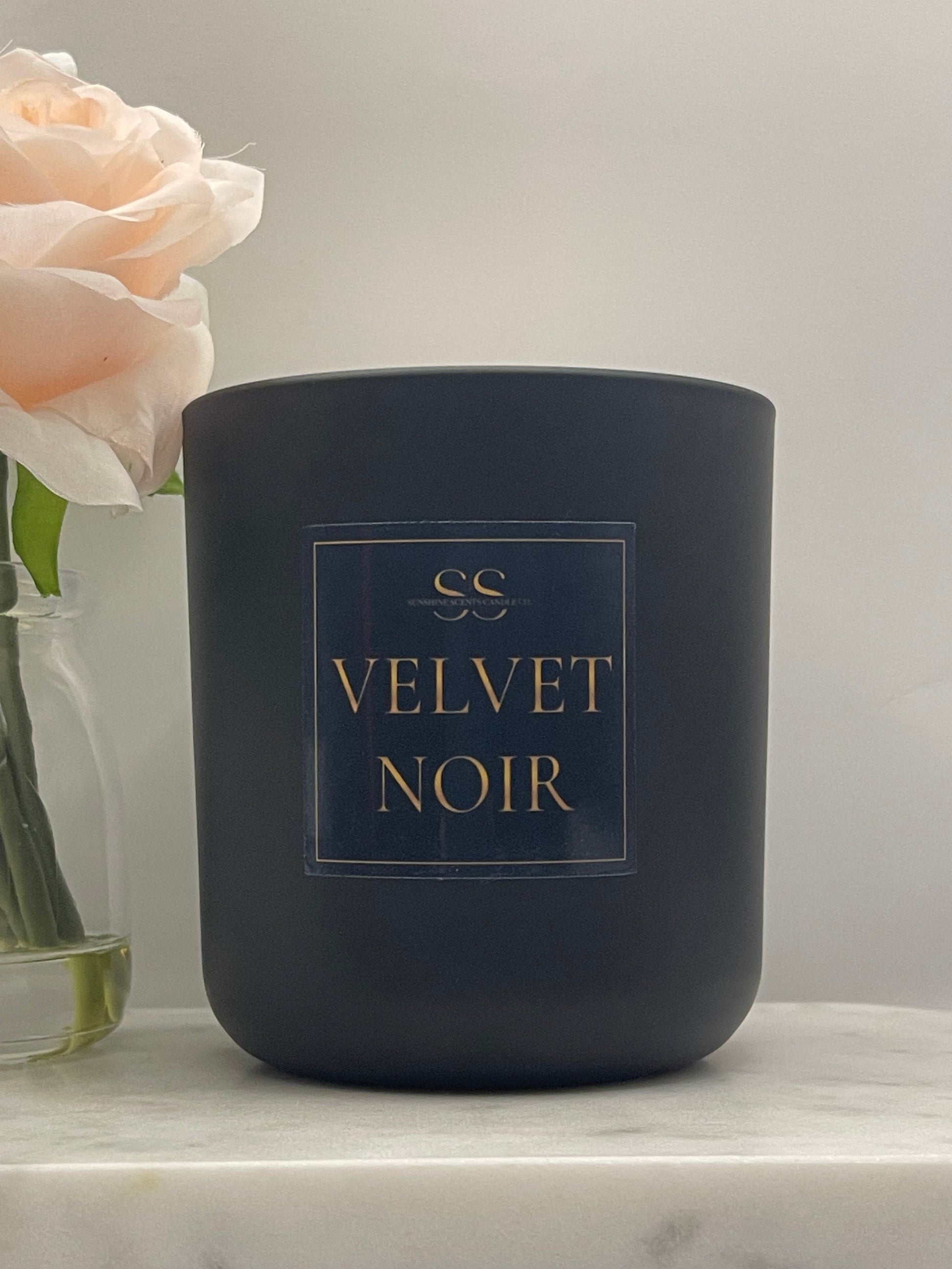 A matte black candle jar with a gold and navy label reading 'VELVET NOIR' and a small monogram logo, photographed on a light marble surface with pale pink flowers in the foreground.