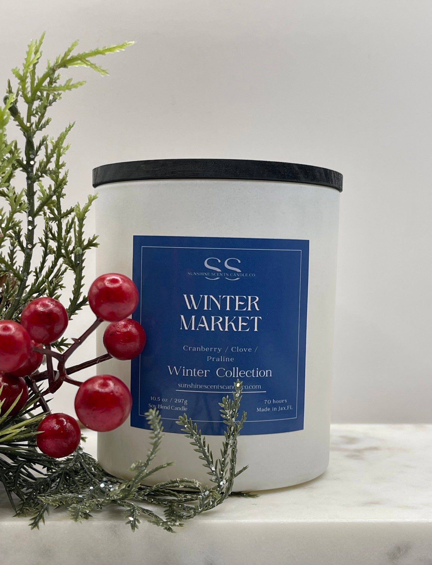 A white candle jar with a black lid and a blue label reading 'Winter Market' and 'Winter Collection,' styled with evergreen branches and red berries on a light marble surface against a soft light gray background.